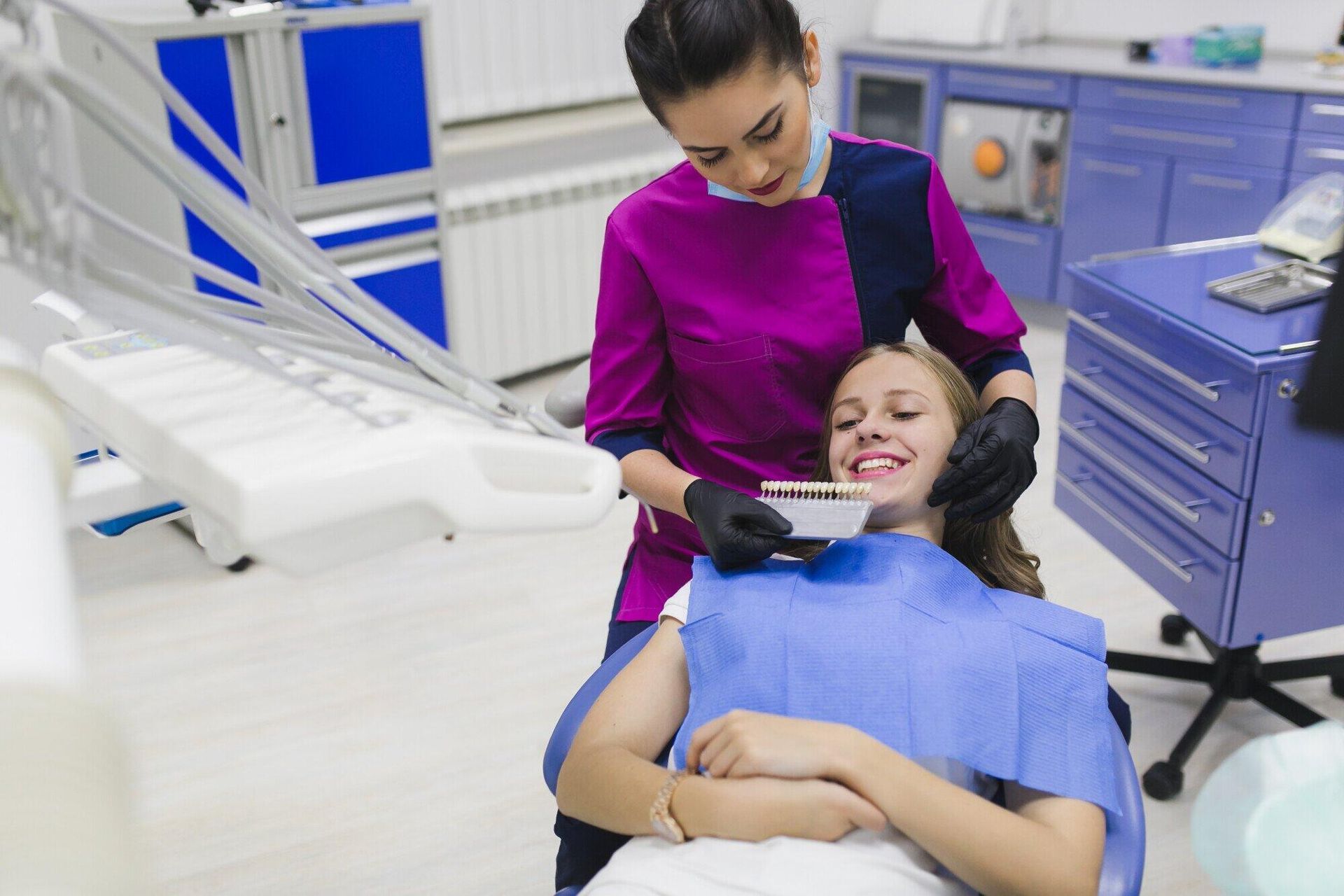 A dental professional in a magenta uniform compares a tooth shade guide to a patient's teeth in a clinical office.