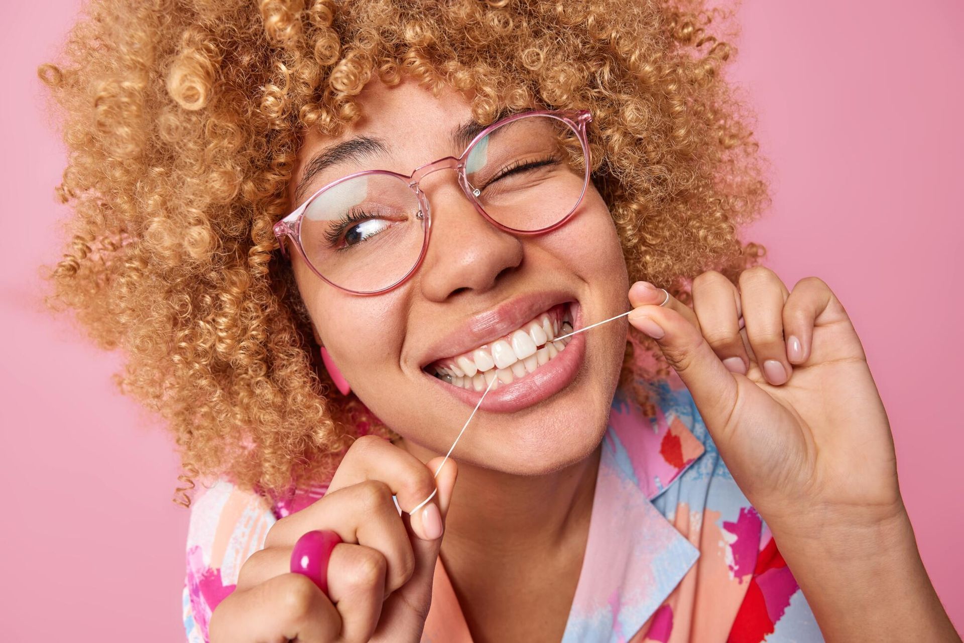 A woman wearing glasses is flossing her teeth on a pink background.