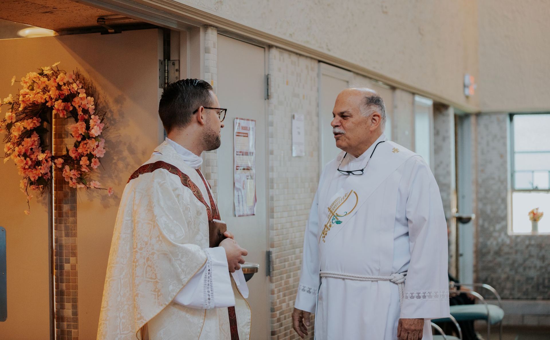 Two clergy in white robes speaking indoors beside a floral wreath display.