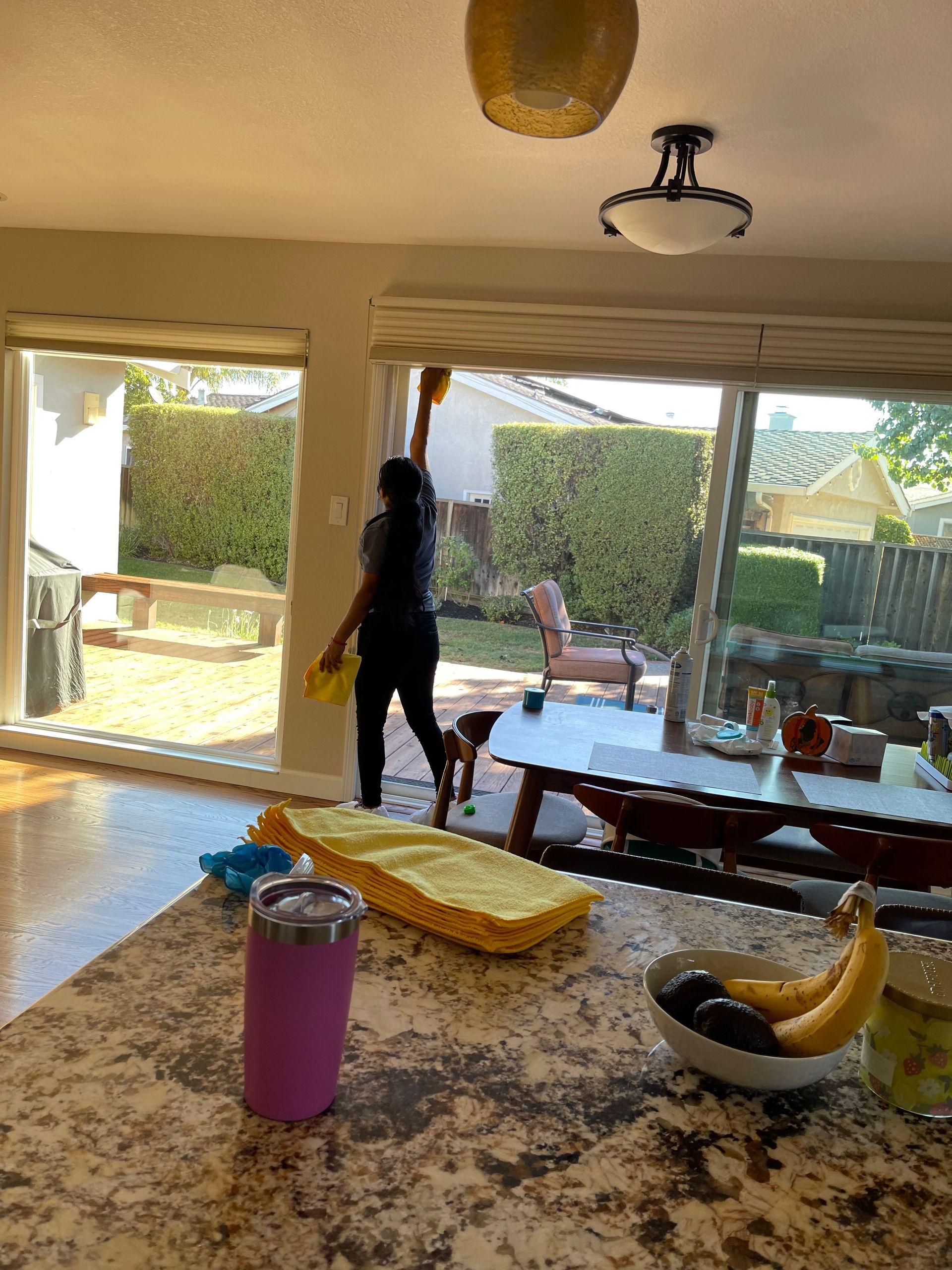 A person uses a duster on a sliding glass door in a brightly lit kitchen with a granite island and bowl of bananas.