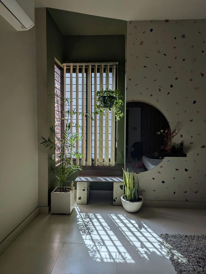 A sunlit corner featuring a window with vertical blinds, potted plants, a small stone bench, and a terrazzo accent wall.