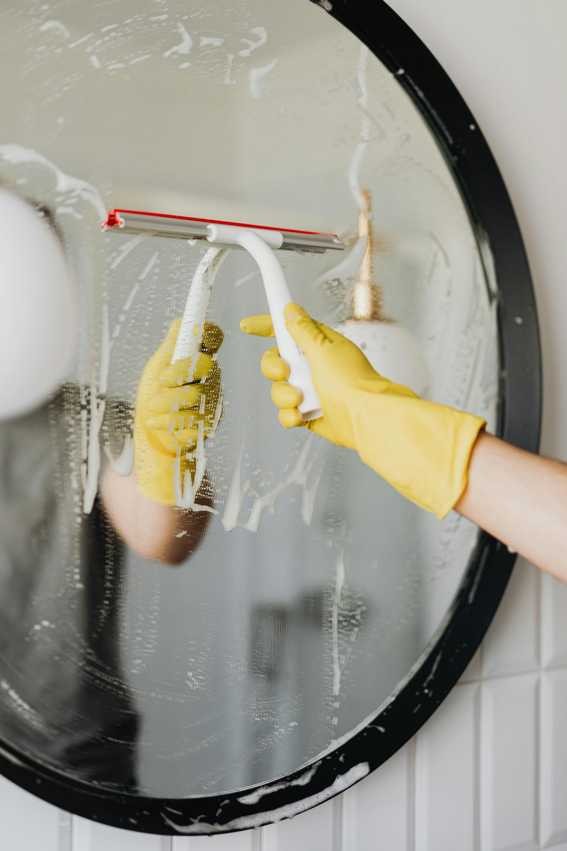 A person wearing yellow rubber gloves uses a squeegee to clean a round mirror.