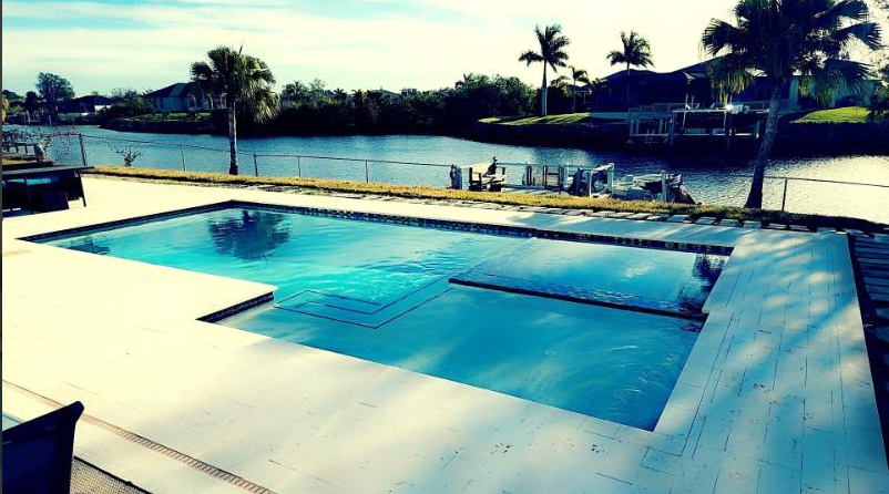 A large swimming pool is surrounded by palm trees and a body of water.