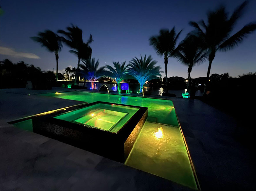 A swimming pool is lit up at night with palm trees in the background