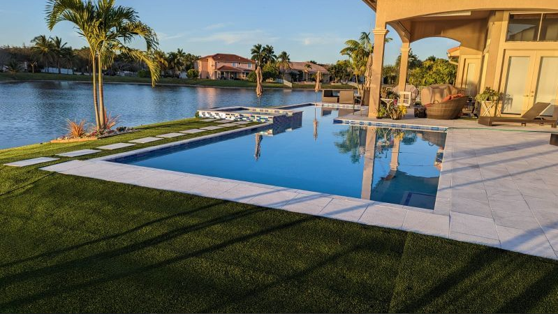 A large swimming pool is surrounded by a lush green lawn next to a lake.