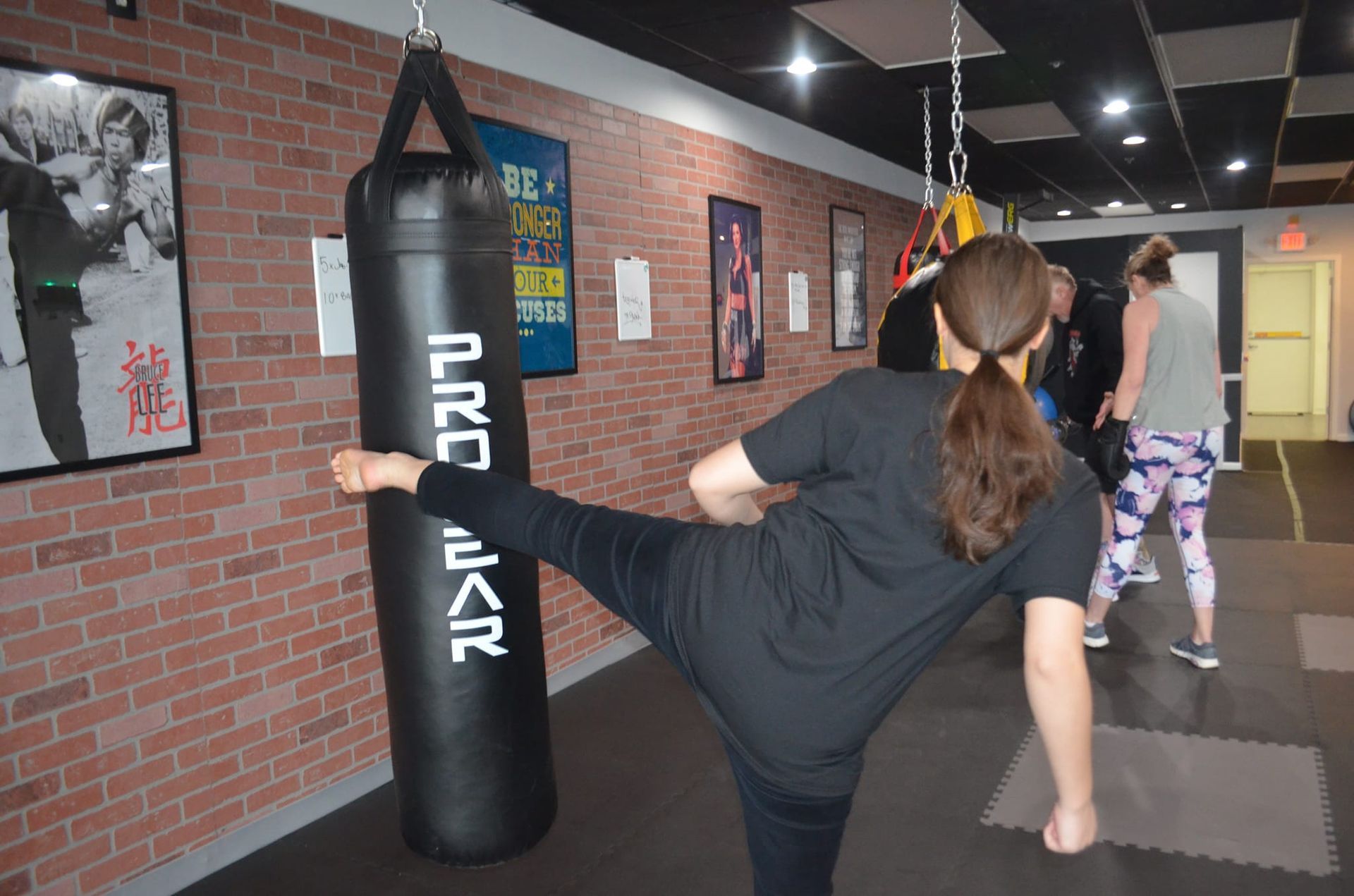 Woman kicks a heavy bag in a gym, brick wall in background.