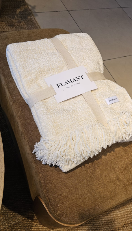 Una coperta pieghevole color crema con trama a frange, legata con una fascia con logo, è appoggiata su un divano marrone.