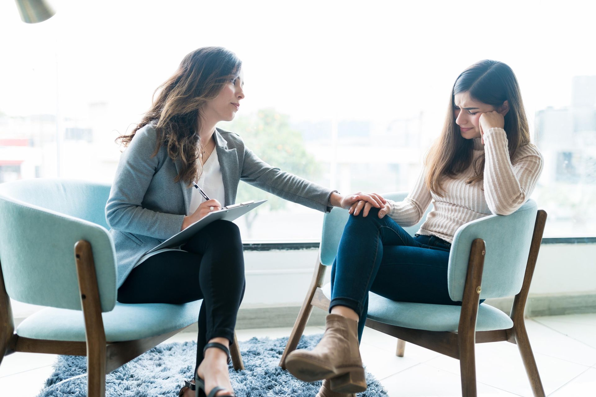 Woman comforting another woman in a light-filled room; one holds a clipboard, the other is visibly upset, both seated.