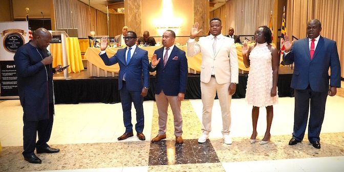 Group of people raising their hands in an oath-taking ceremony. A man in a blue suit speaks on the left.