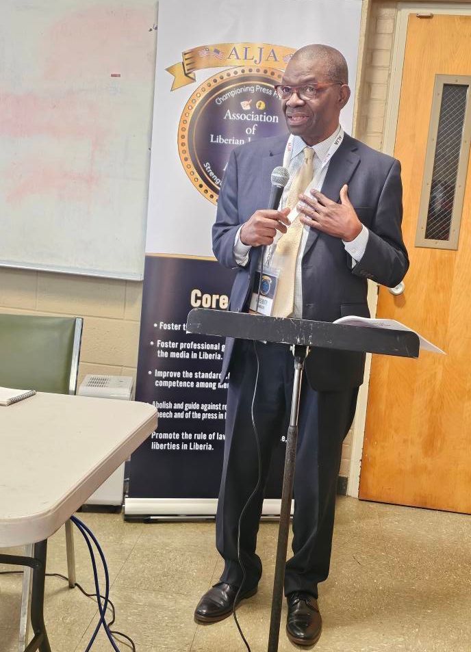 James Butty in suit speaking at a lectern, holding a microphone, with a banner in the background.