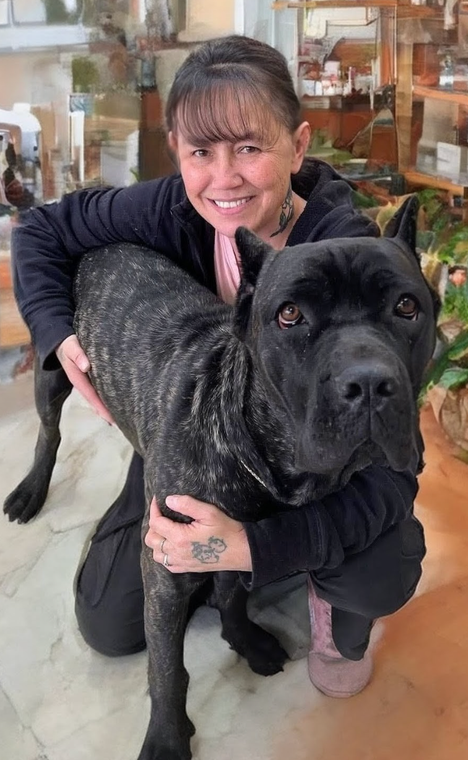 Woman kneeling, hugging large brindle dog. Both smiling at camera. Indoors, plants in background.