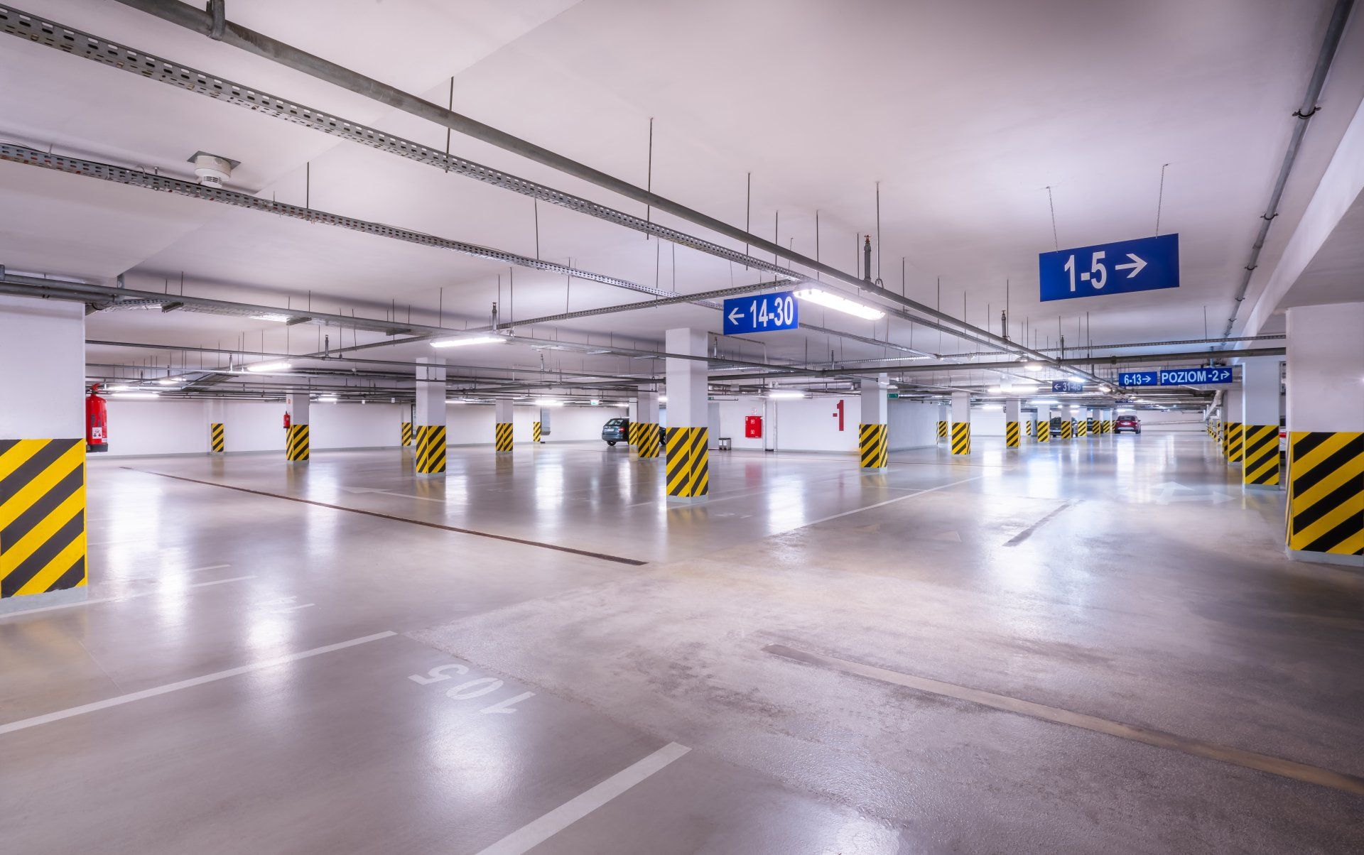 Pusty, jasno oświetlony parking wielopoziomowy z żółto-czarnymi słupami wsporczymi. Znaki prowadzą na poziomy parkingowe.