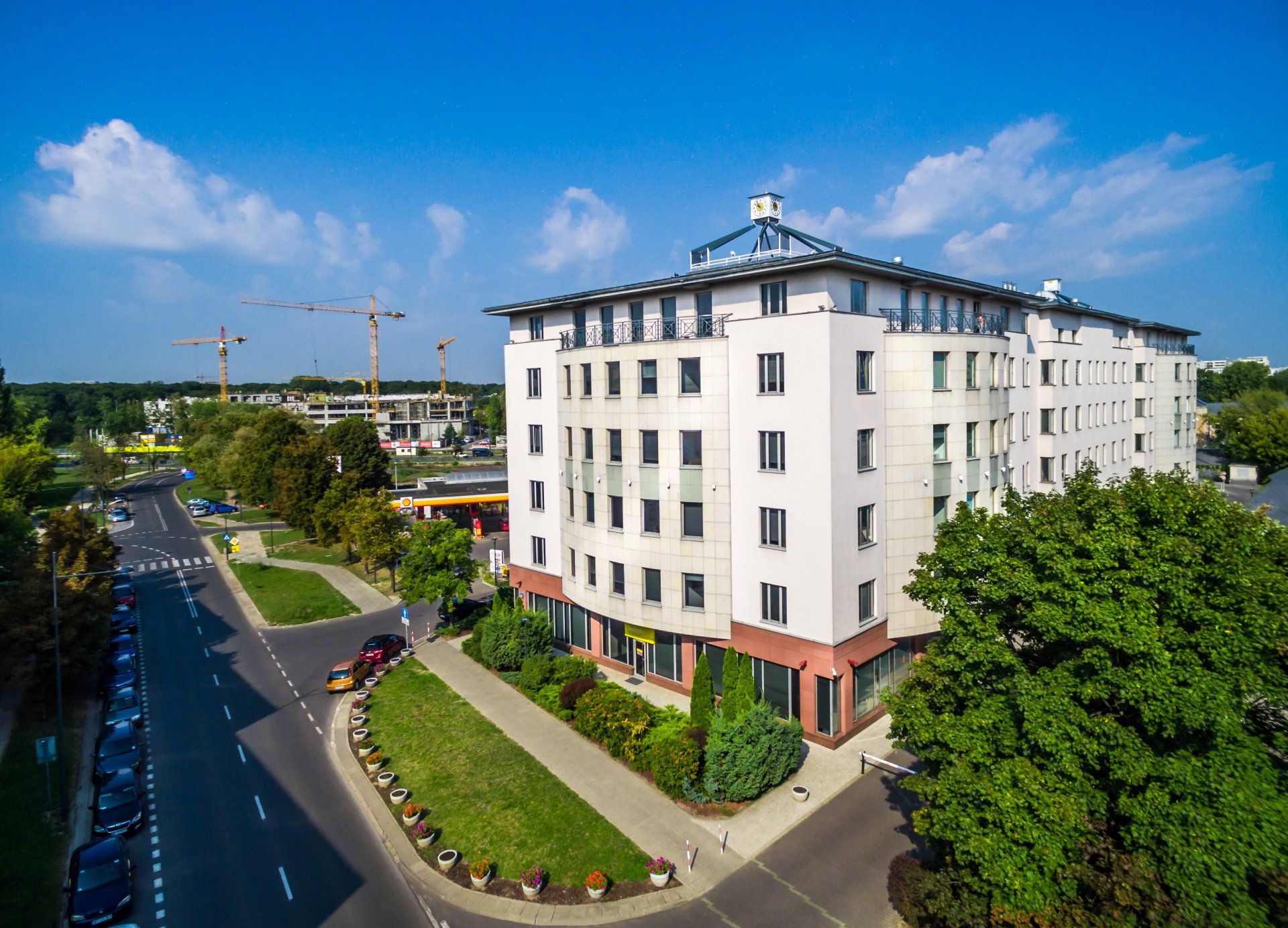 Wielopiętrowy biały budynek ze szklanym dachem, widok na ulicę. Widoczne drzewa i dźwigi budowlane.