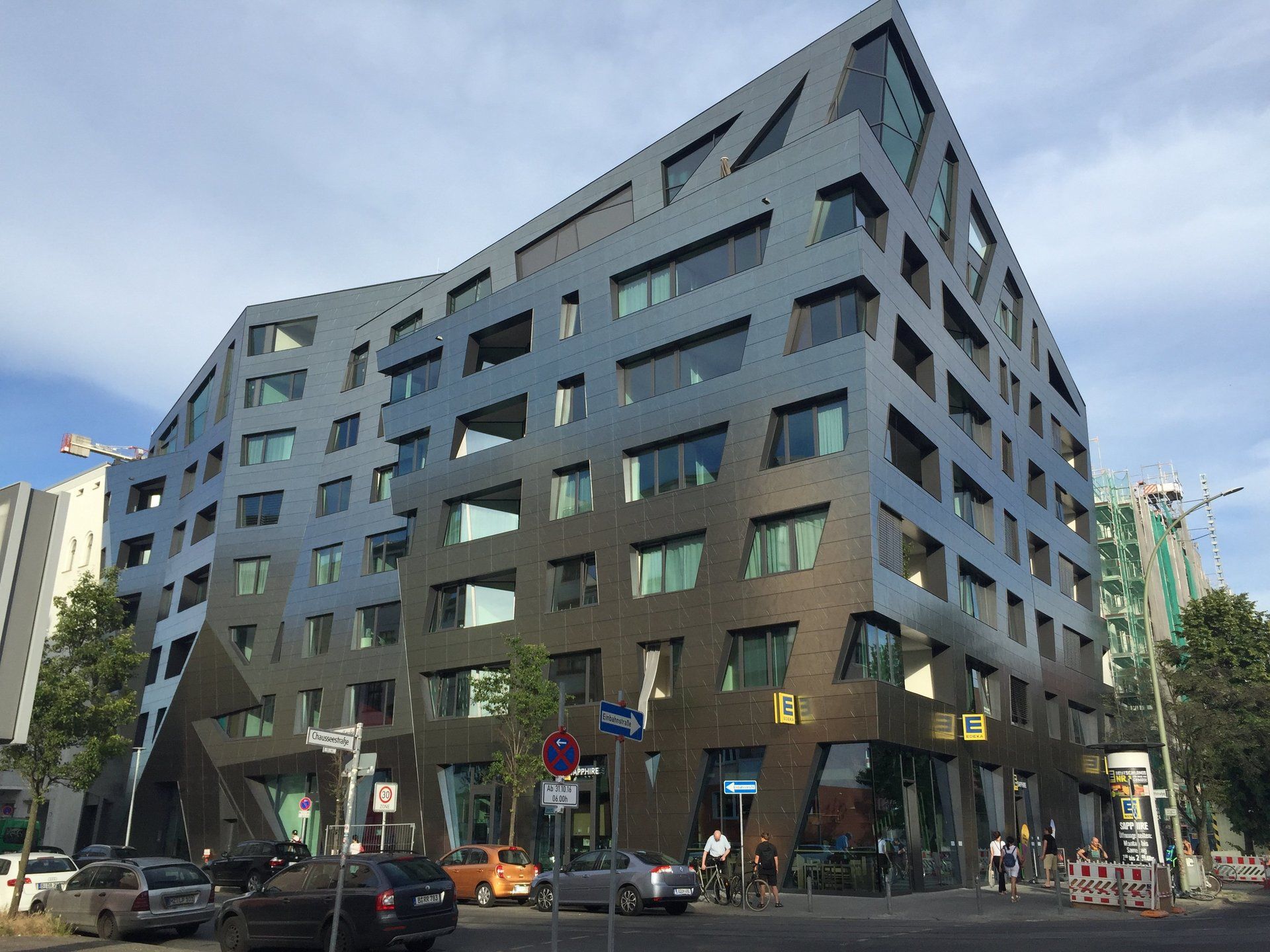 Modern angular building with dark facade, street signs, and parked cars. Blue sky.