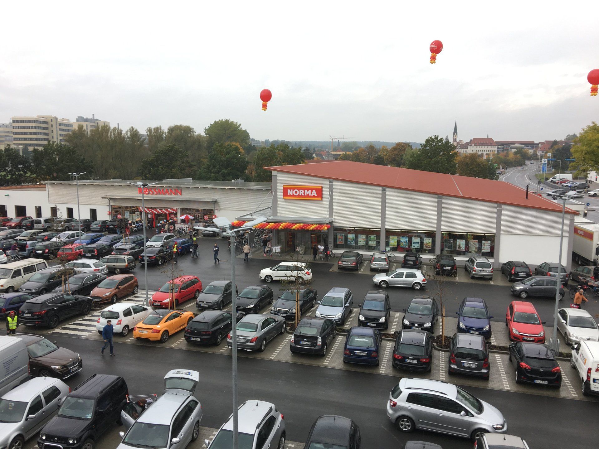 Busy parking lot in front of a supermarket; red balloons in the sky.