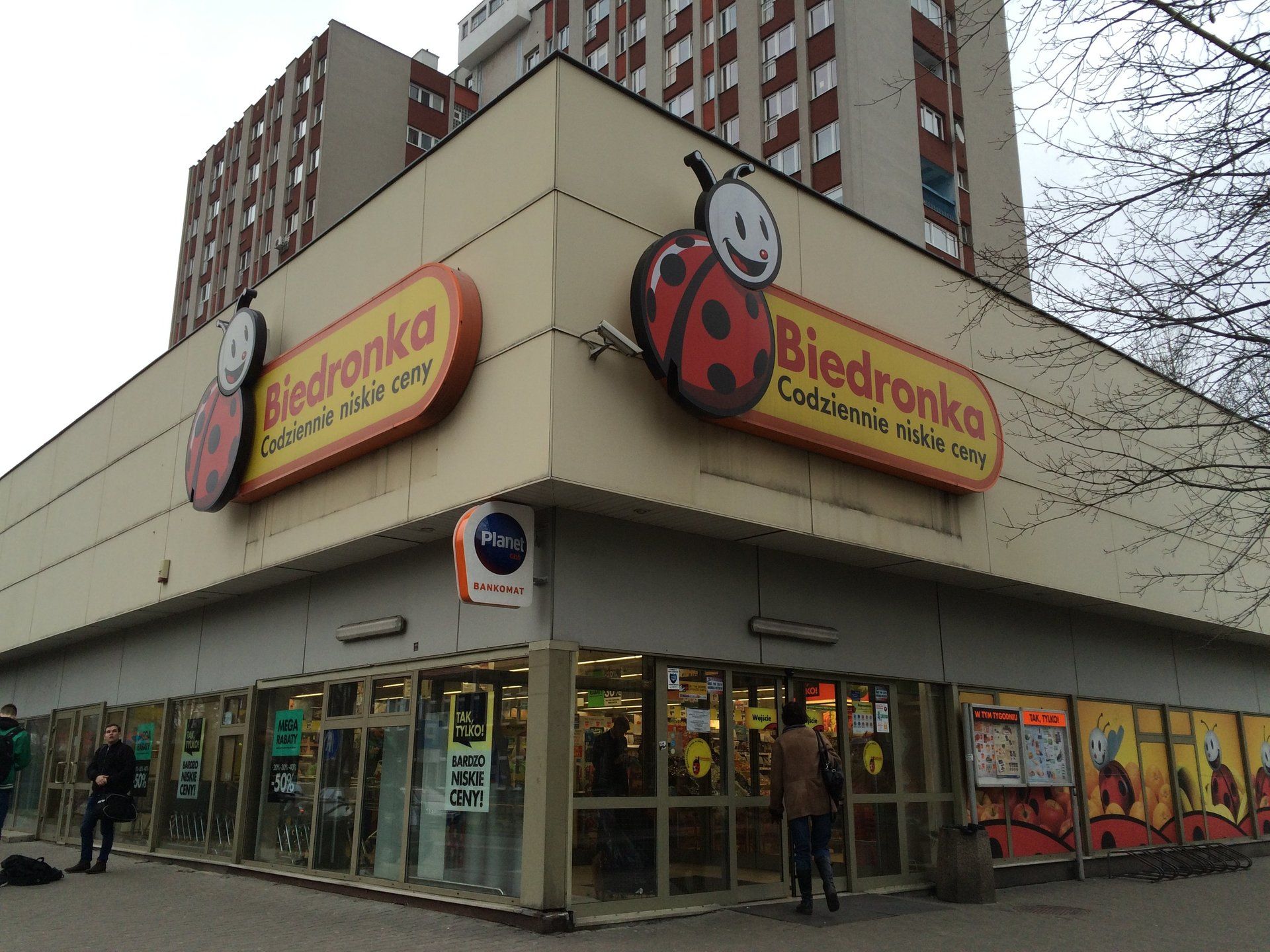 Biedronka grocery store with ladybug logo on a building corner. People stand outside. Brown and beige tones.