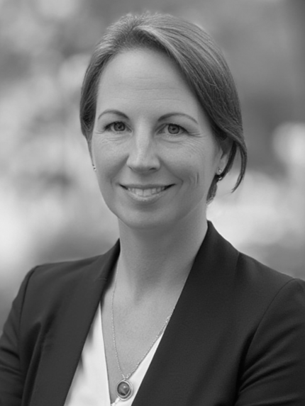 Frau in Blazer und gepunktetem Hemd, leicht lächelnd, vor einer Stadtkulisse.