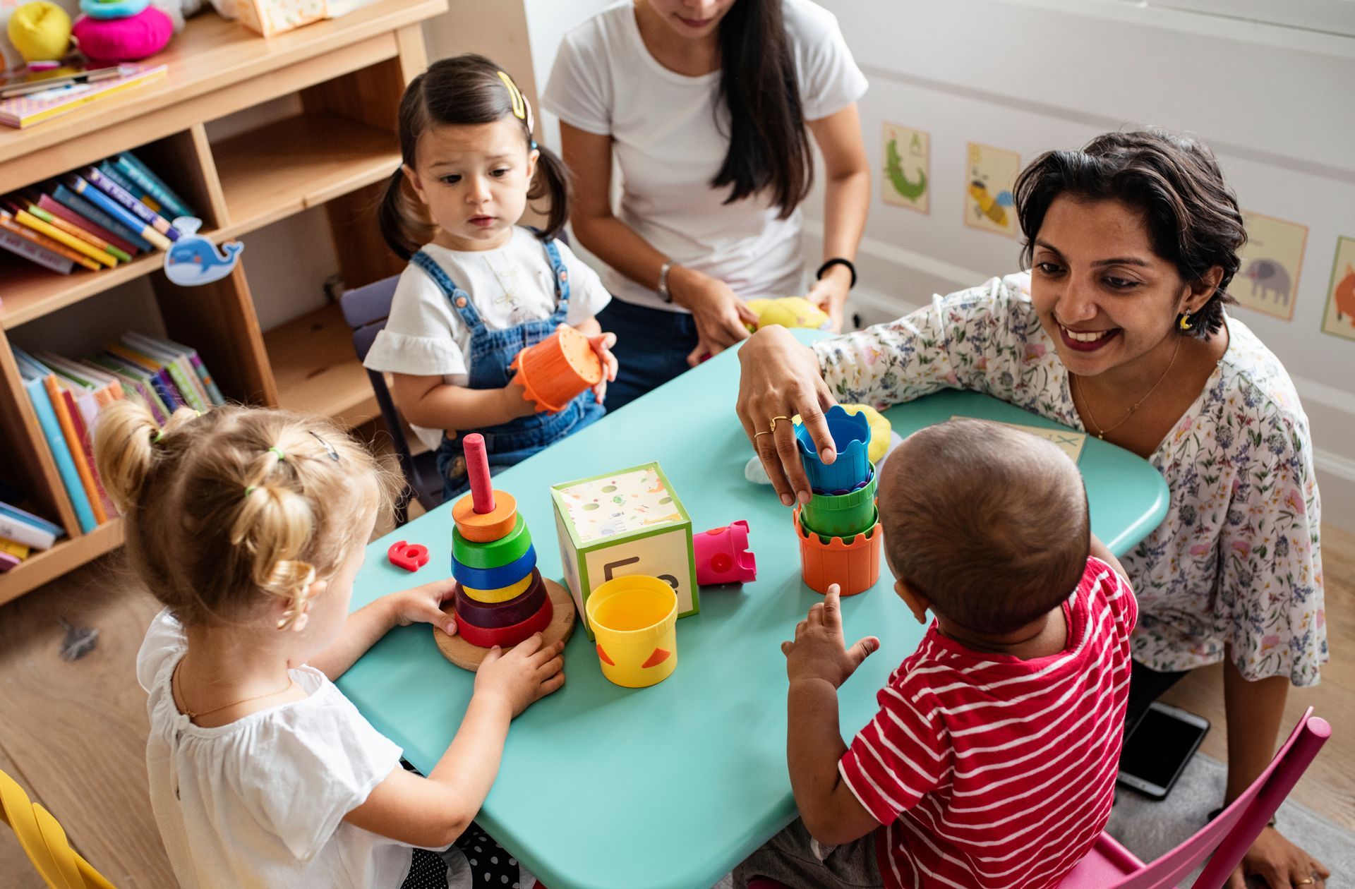 Children playing with colorful stacking toys at a daycare table with two adults assisting.