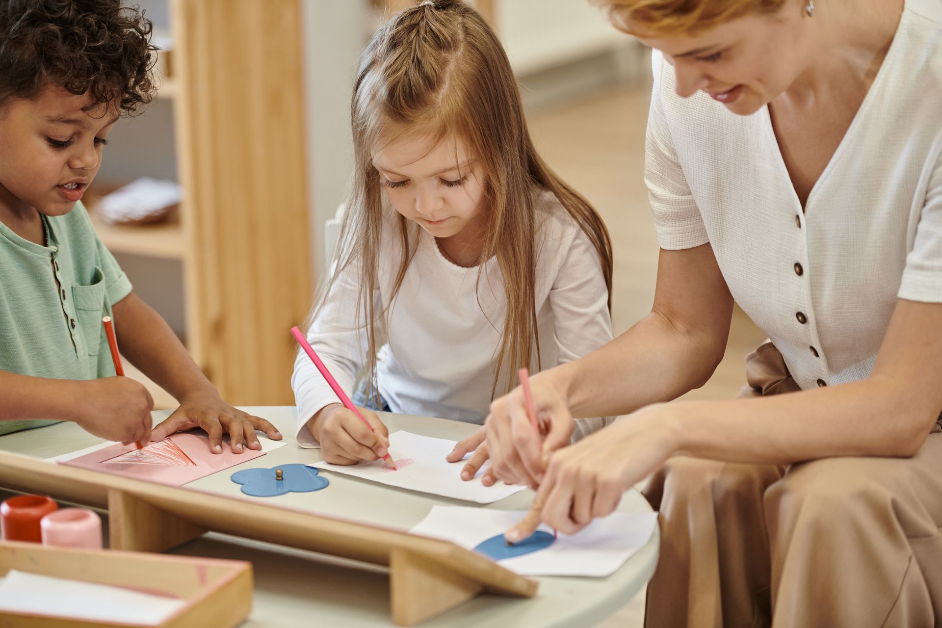Two kids are drawing on a paper while a teacher watches them.