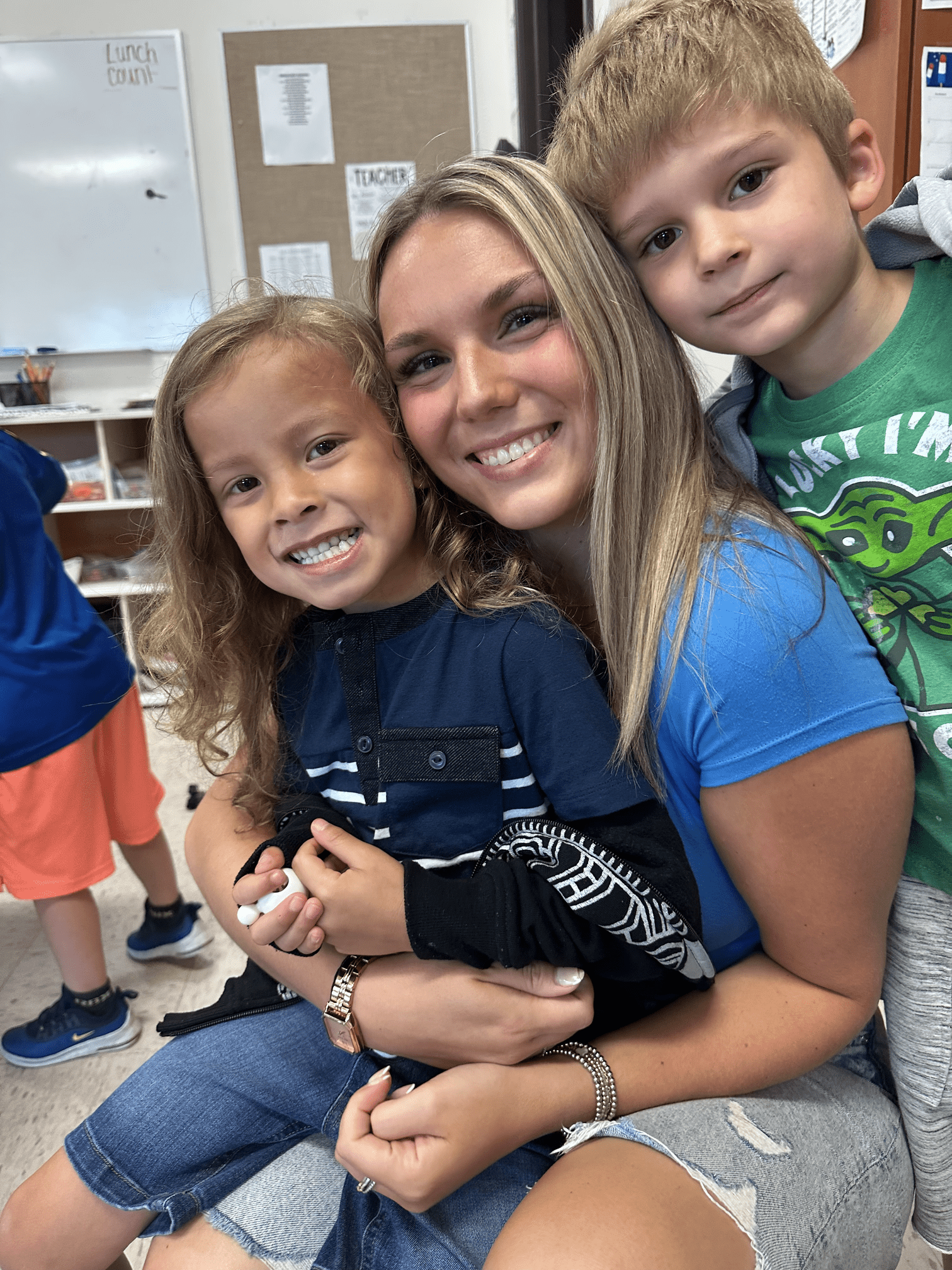 Woman smiles hugging two young children in a classroom.
