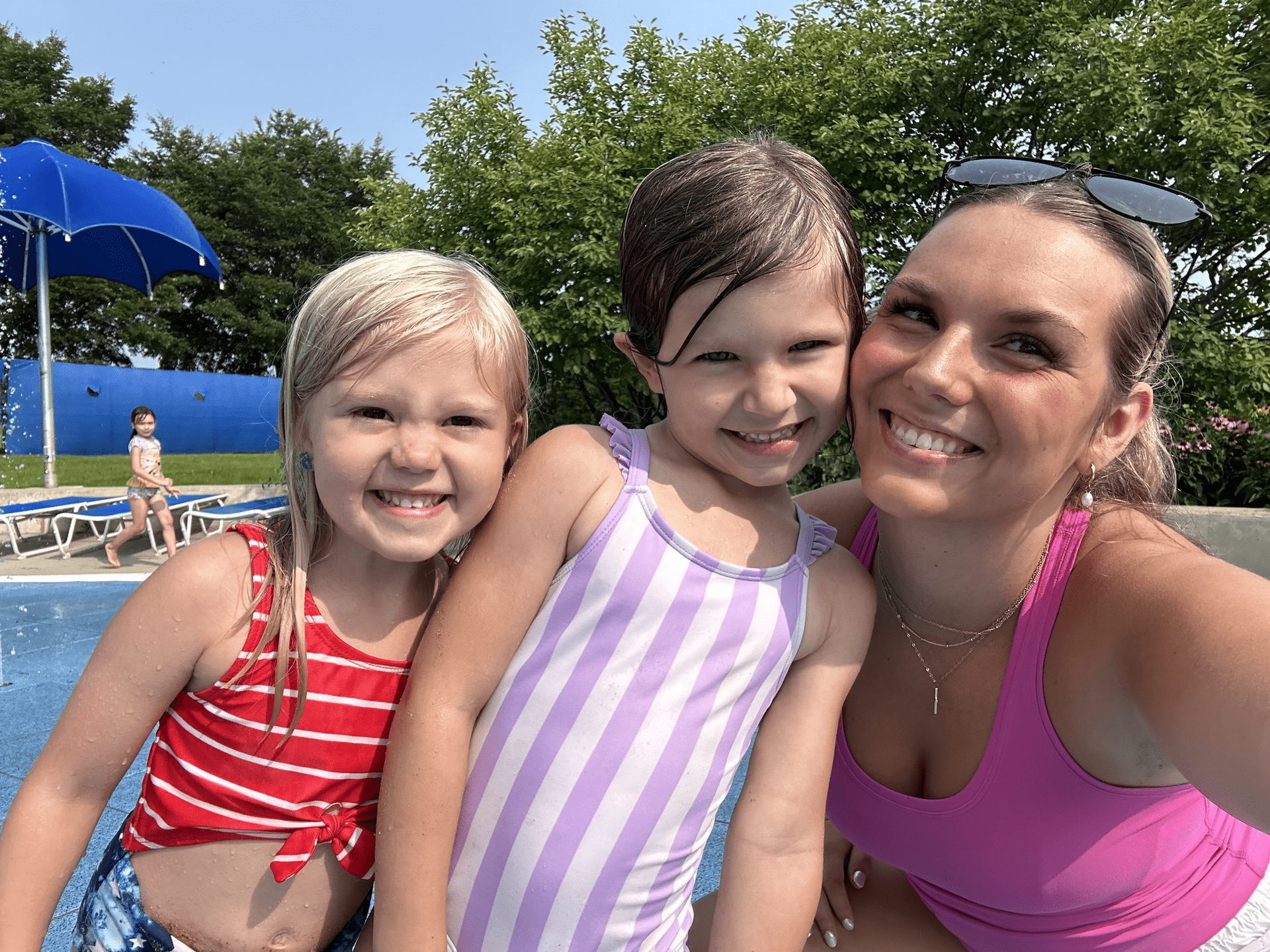 Woman and two young girls smiling at a pool. One girl wears red and white, the other purple stripes.