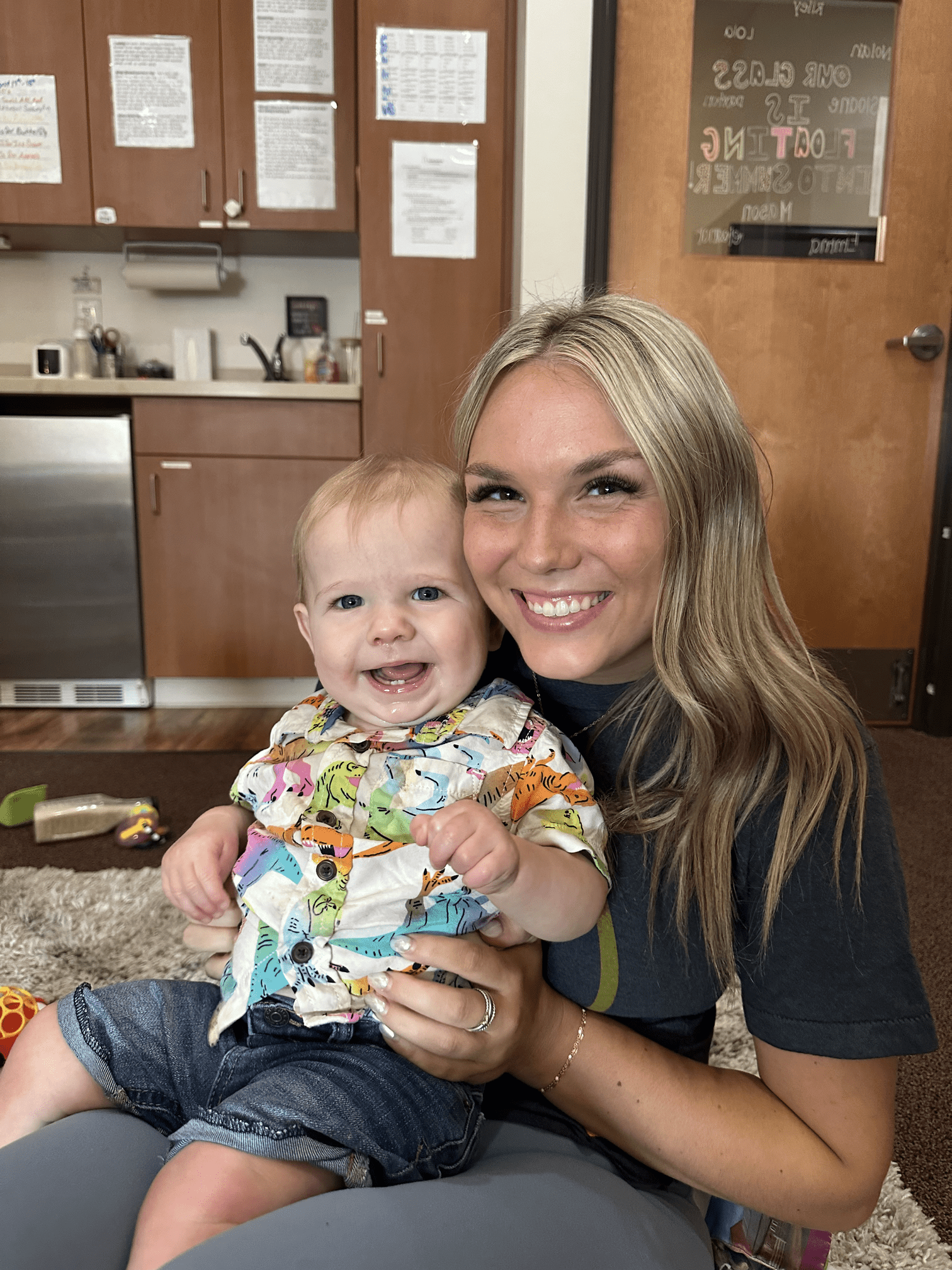 Smiling woman holding a laughing baby; both sitting on a floor.