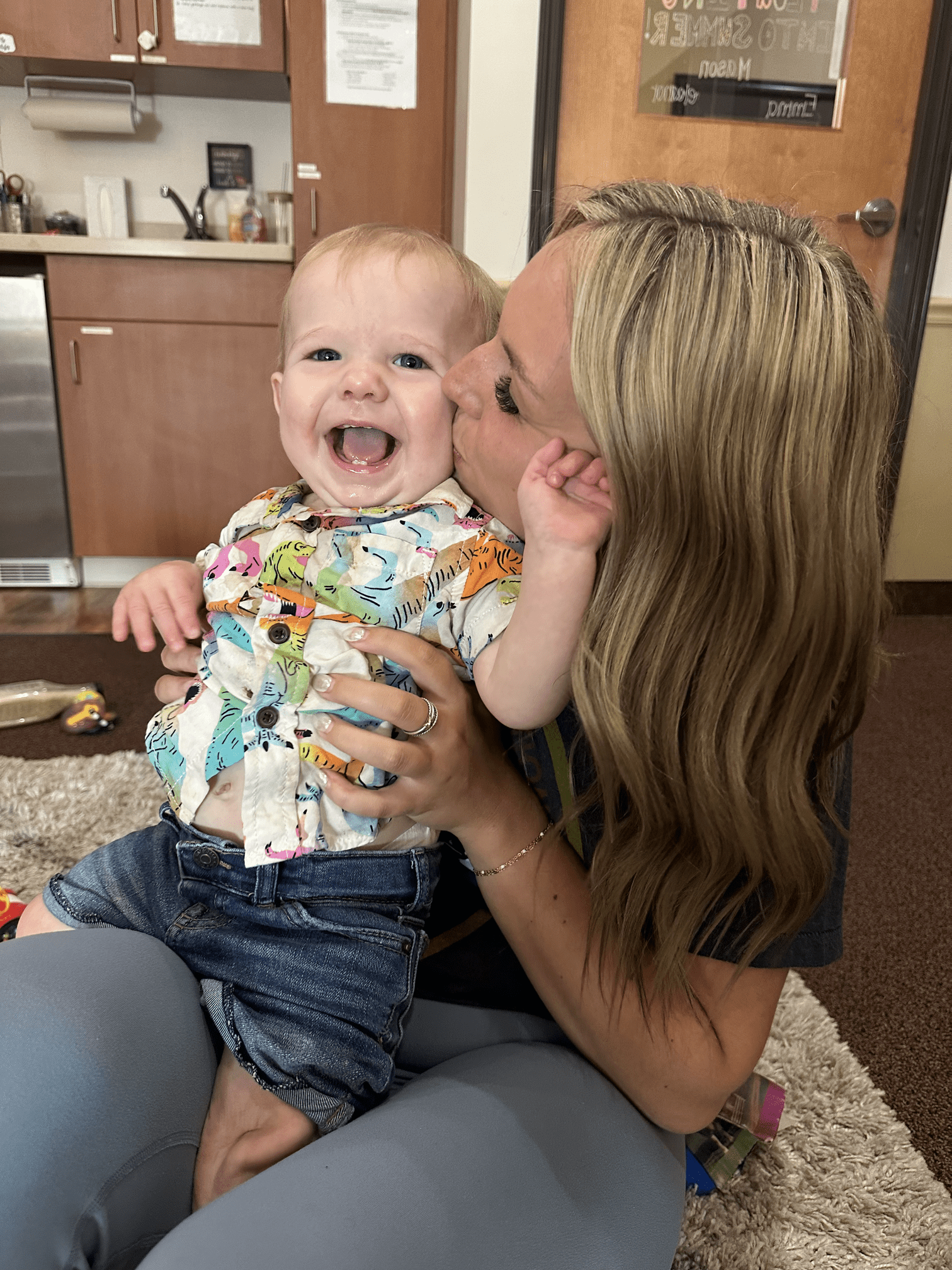 Woman kisses a laughing baby on the cheek indoors. Baby wears a colorful shirt and jean shorts.
