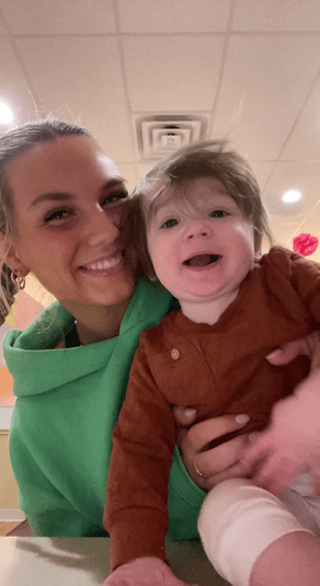 Woman in green hoodie holds a smiling baby in a brown shirt. Inside setting.