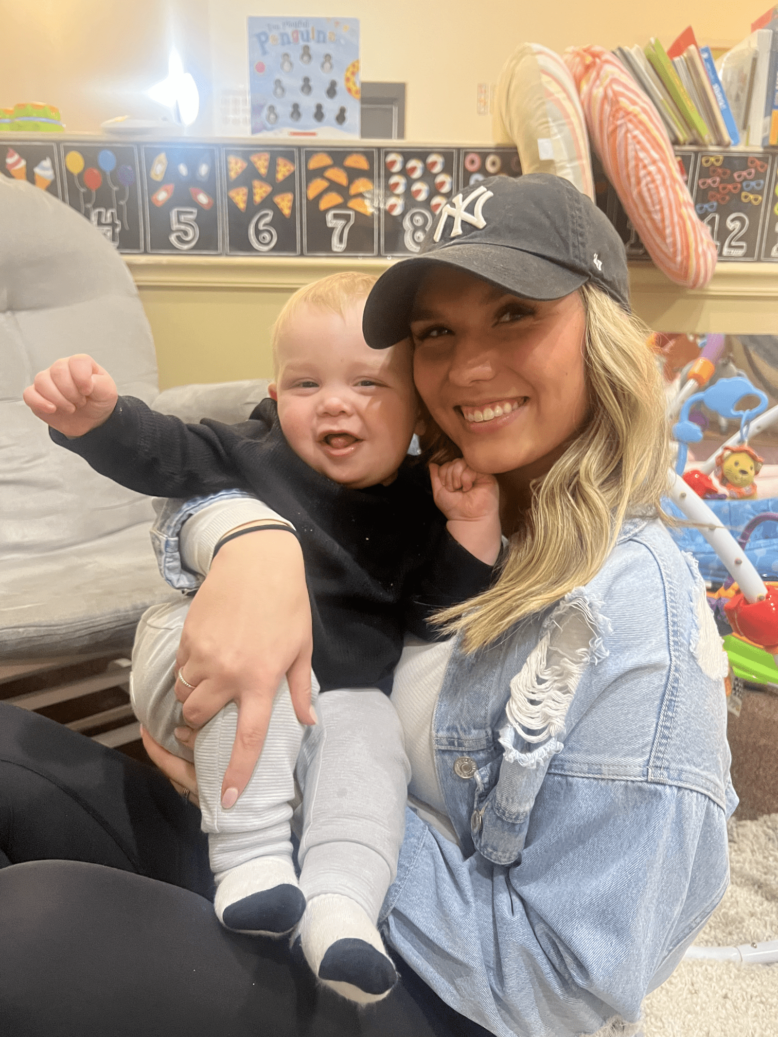 Woman in denim jacket and baseball cap holds a laughing baby indoors, surrounded by toys.