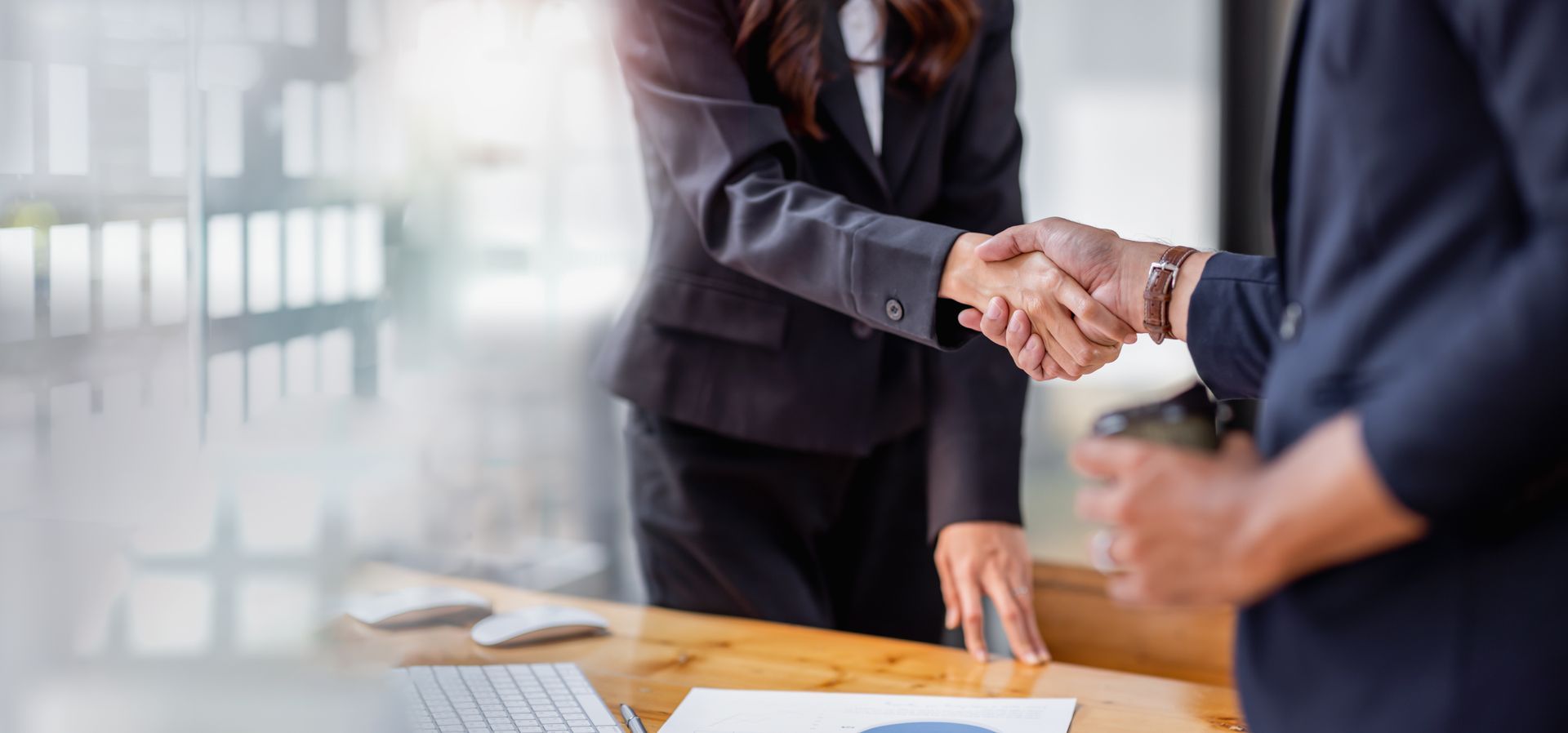 Two professionals in suits shaking hands over a wooden desk with a computer keyboard and documents in an office.