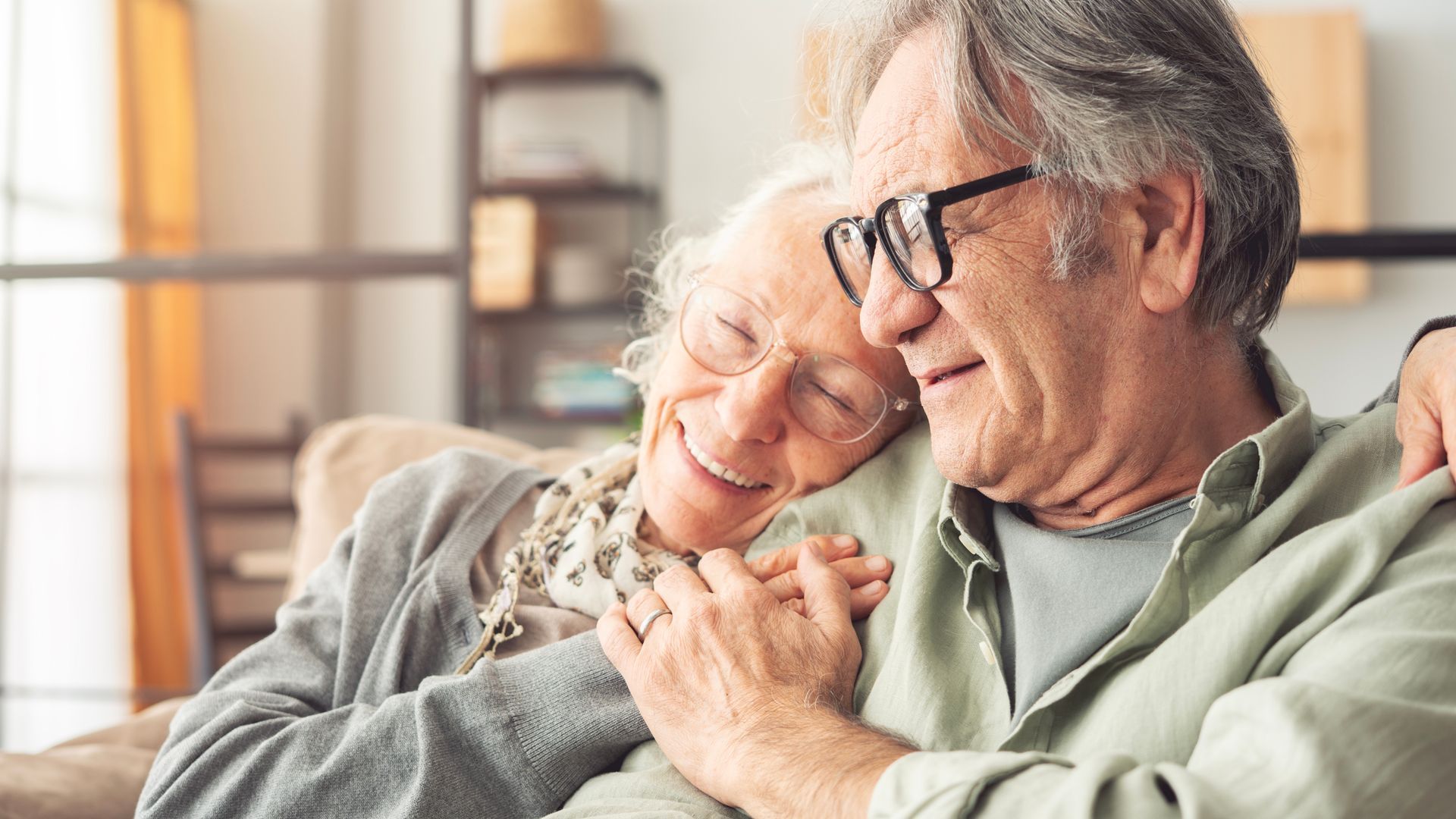 Two people sit closely together, smiling, with one resting their head on the other's shoulder in a warm, indoor setting.