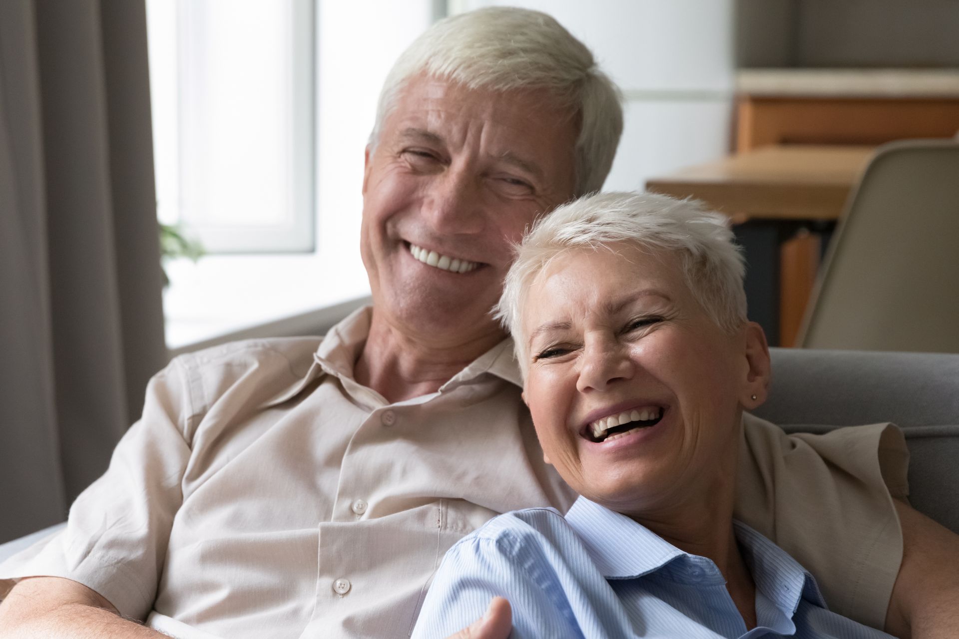 A couple sitting together on a couch, smiling and laughing in a bright, indoor living space.