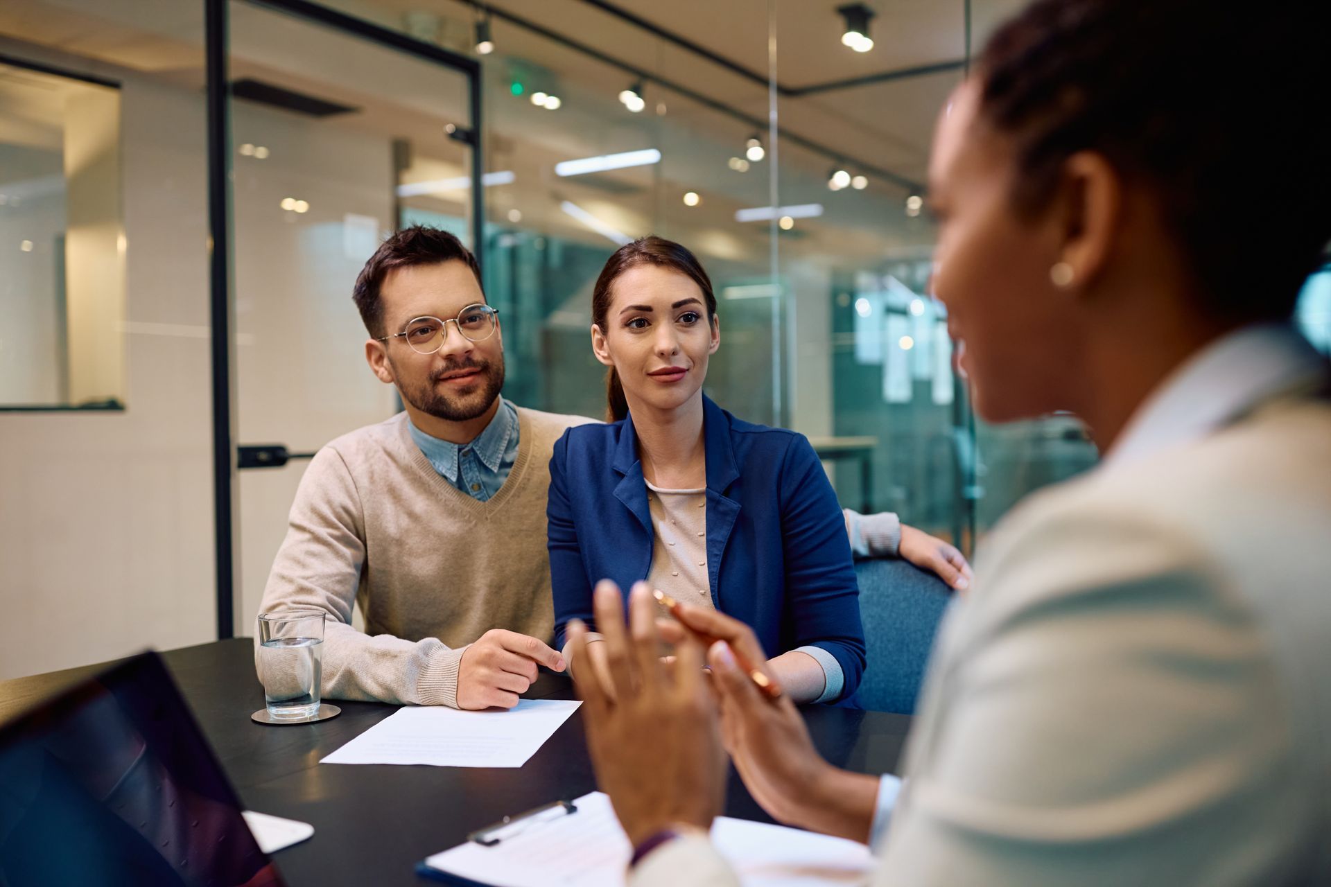A business advisor in a suit discusses documents with a couple sitting at a table in a glass-walled office.