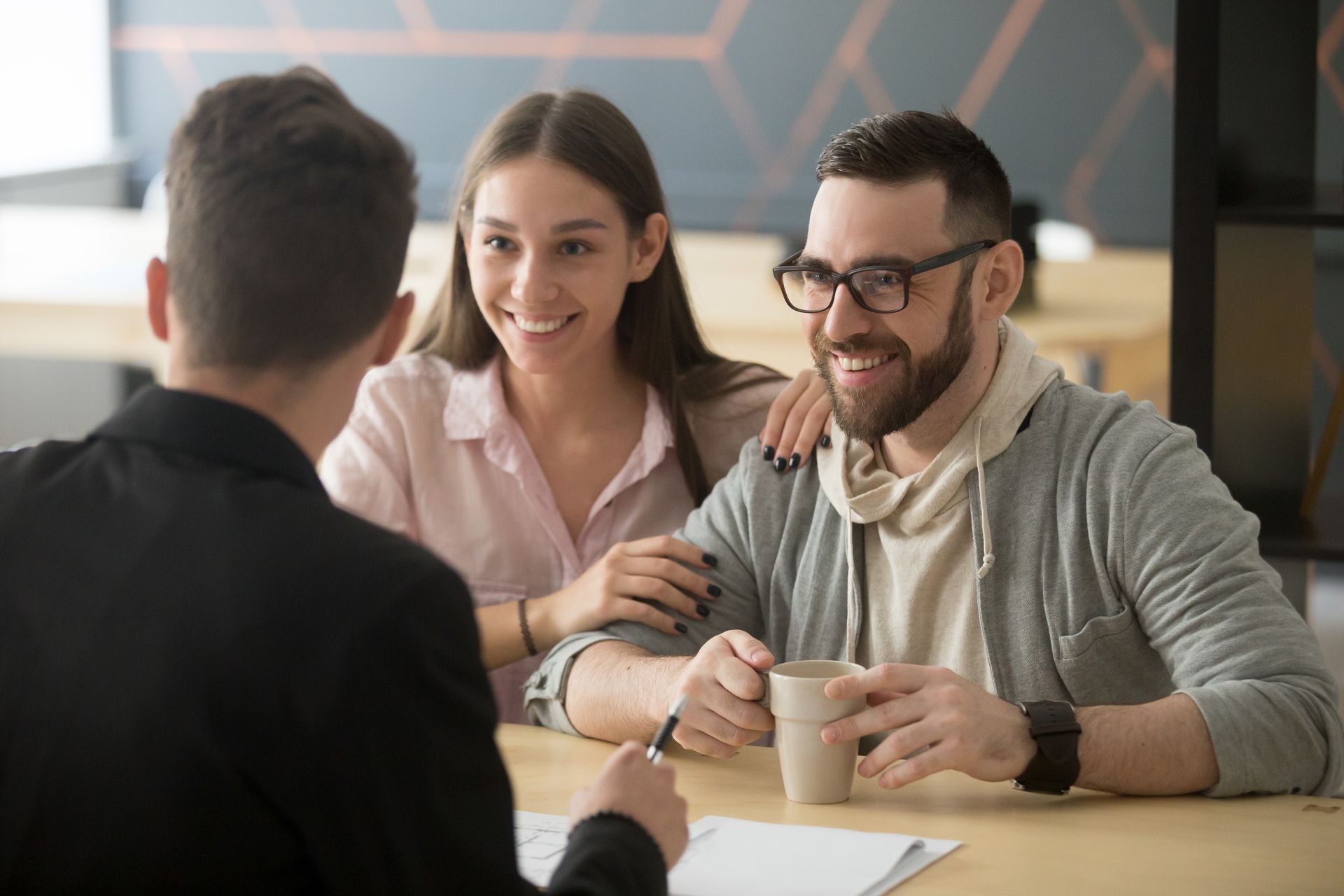 A couple smiles while sitting at a table across from a professional, who is reviewing documents with them.