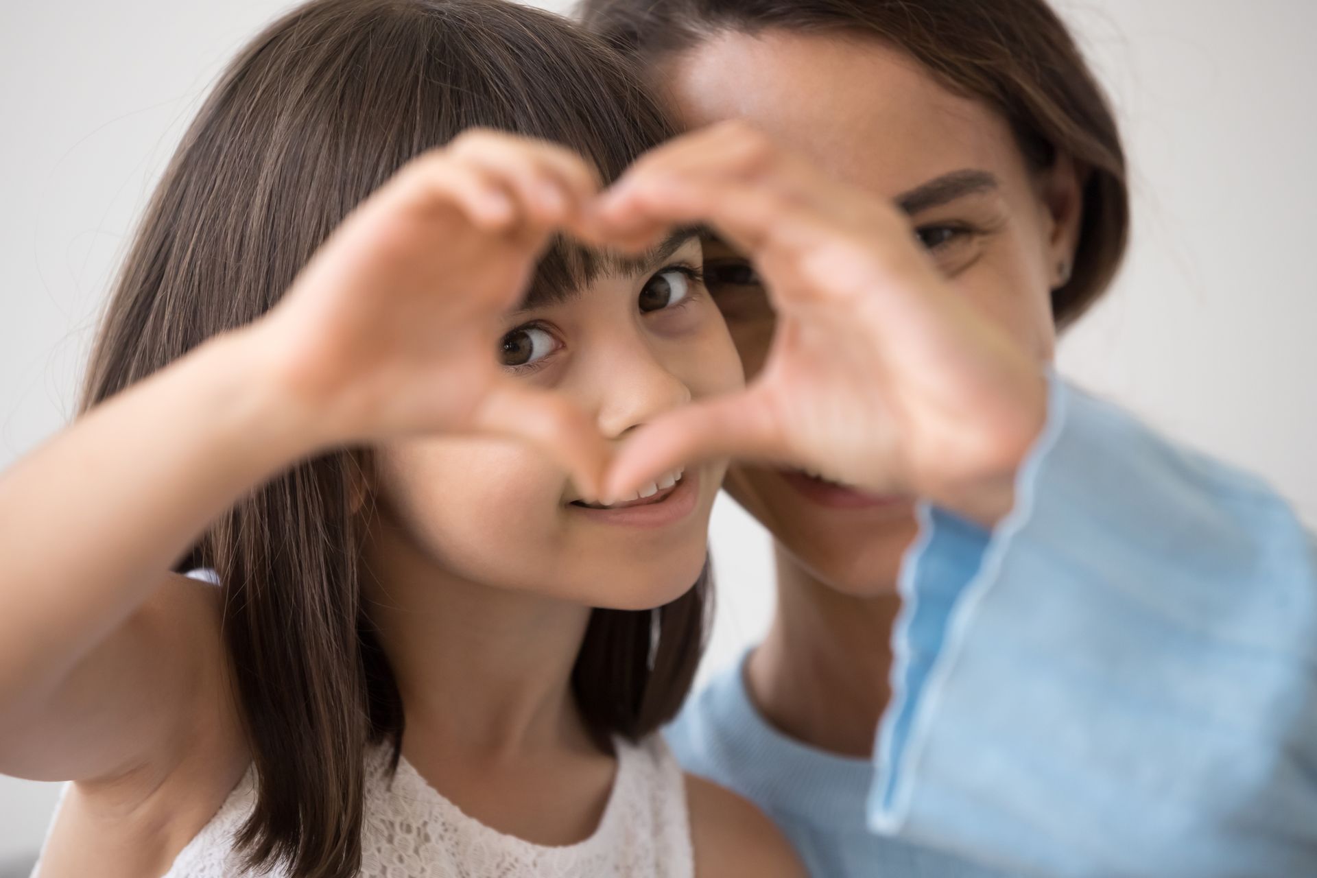 A person and child looking through a heart shape made with their hands, both smiling.