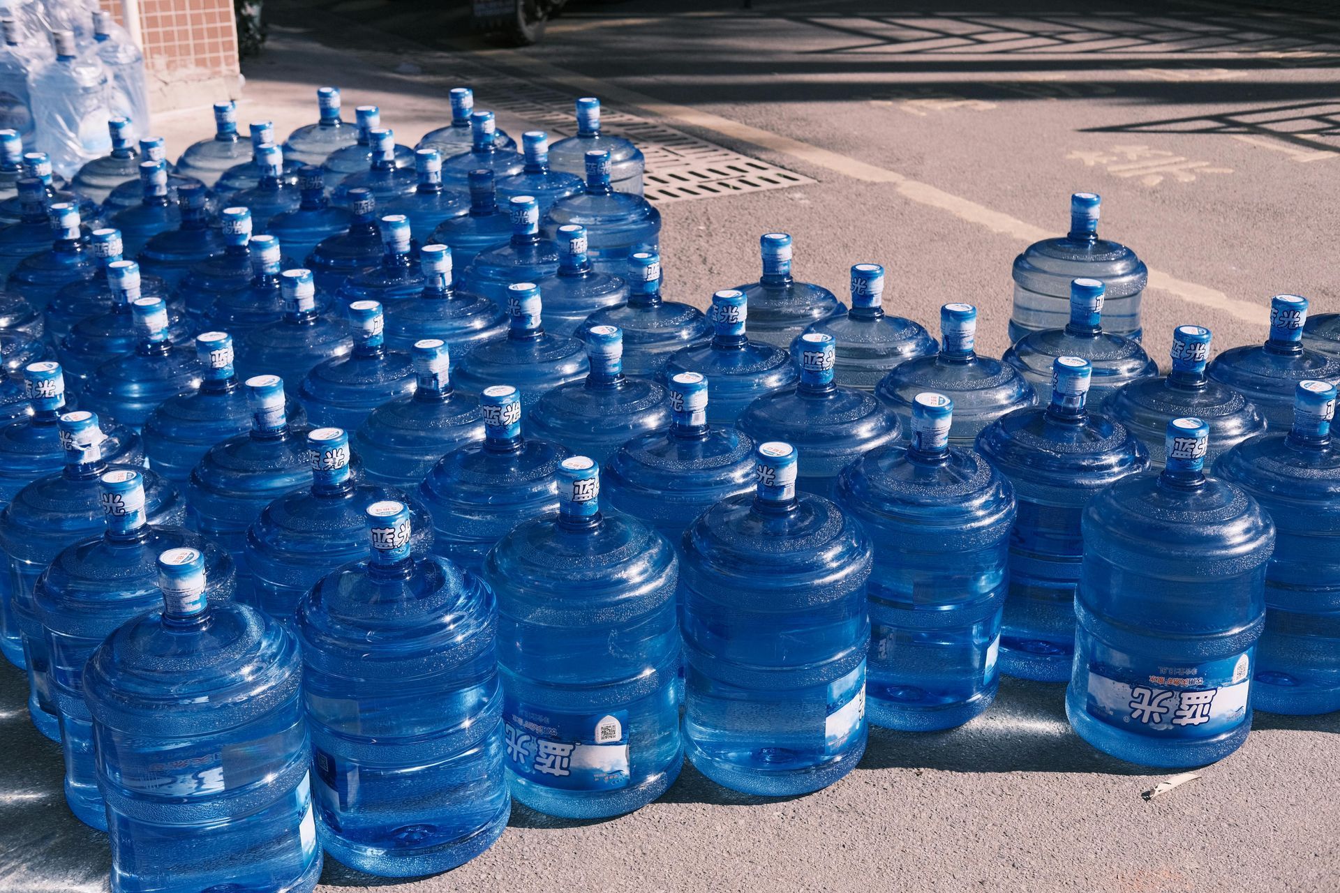 Rows of large, blue water cooler jugs arranged outdoors on a paved surface.