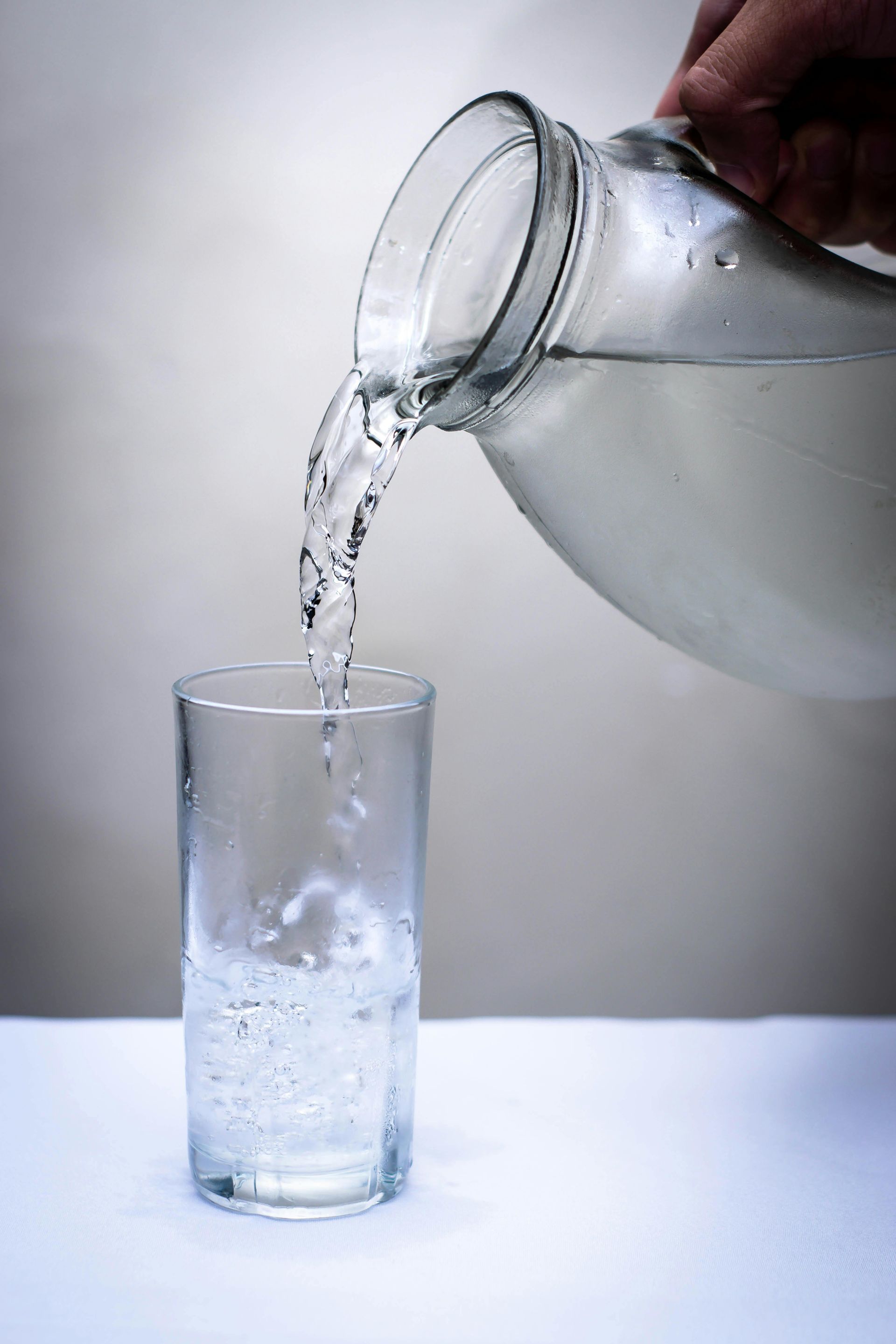 A hand pouring water from a glass pitcher into a tall glass on a white surface.