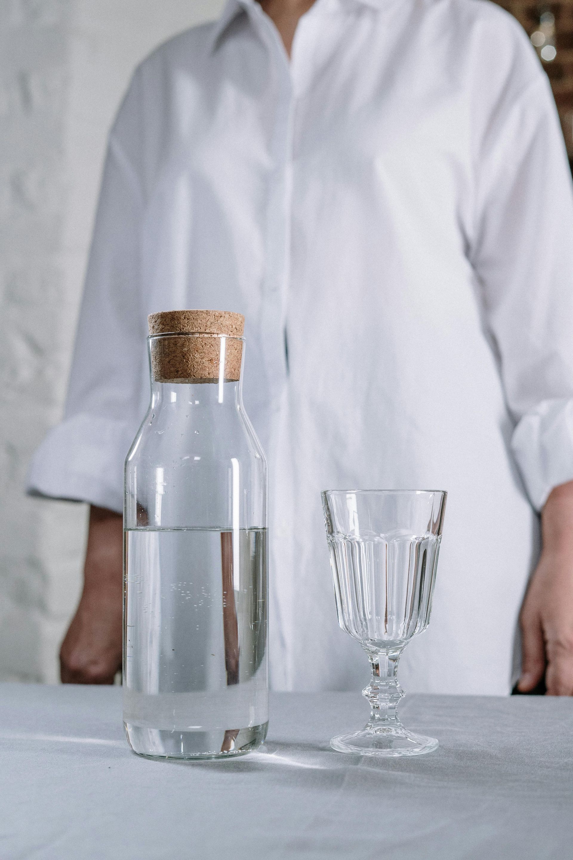 A person in a white shirt stands behind a glass water bottle and a crystal glass on a light-colored surface.