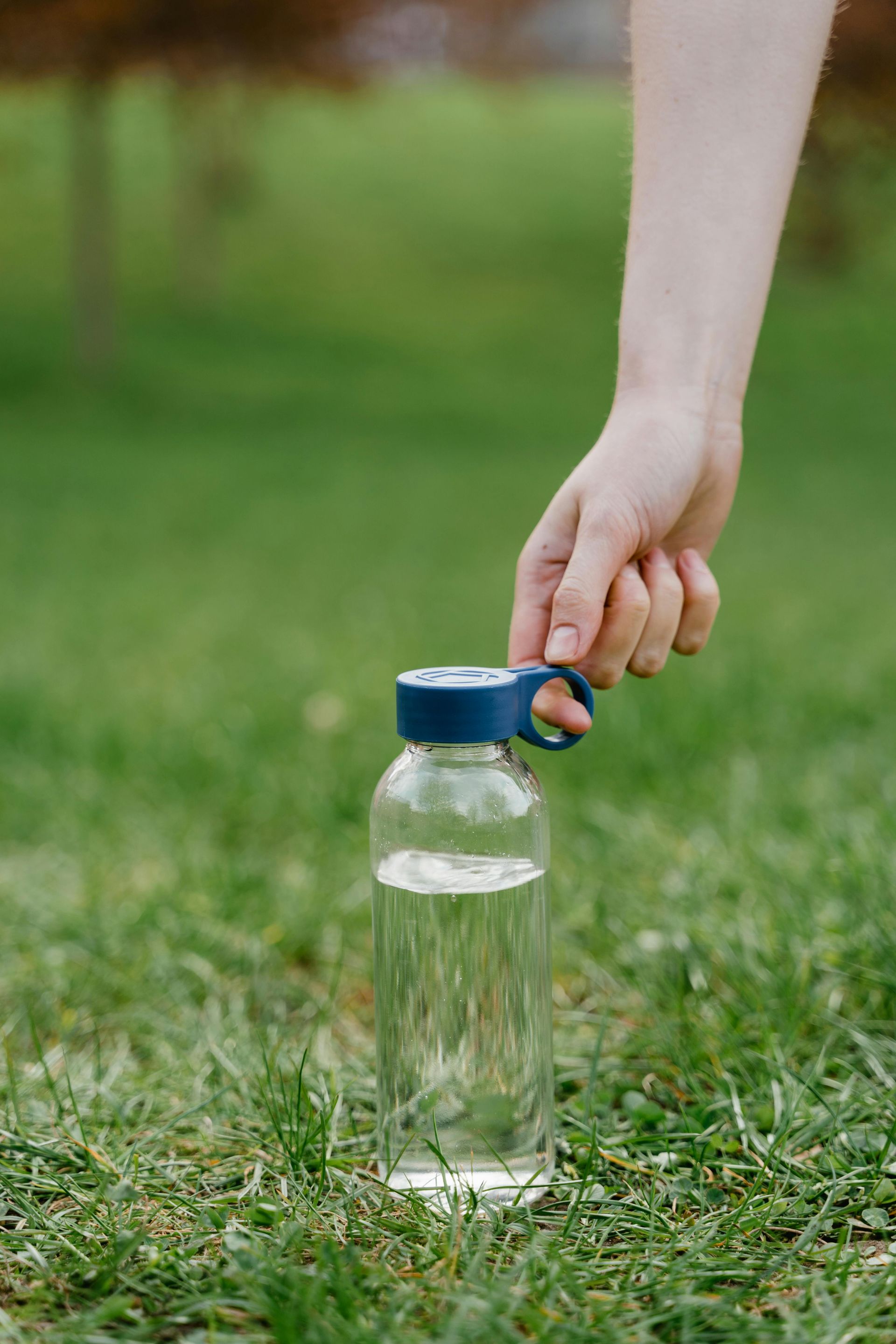 Hand holding a water bottle filled with clear liquid on a grassy lawn. The bottle has a blue cap.