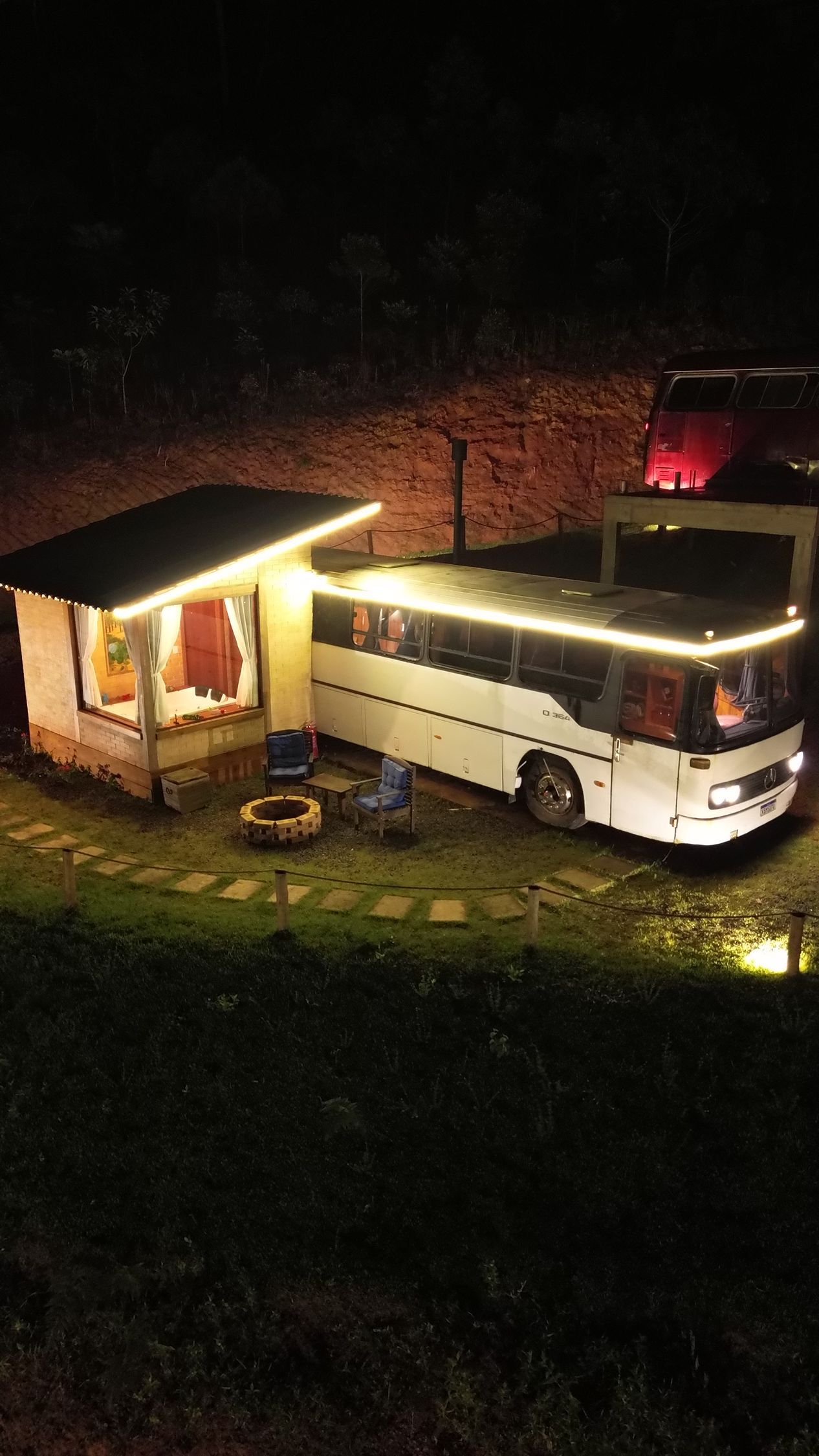 Ônibus transformado em moradia com estrutura anexa, iluminado com luzes de cordão à noite. Villa Caravaggio