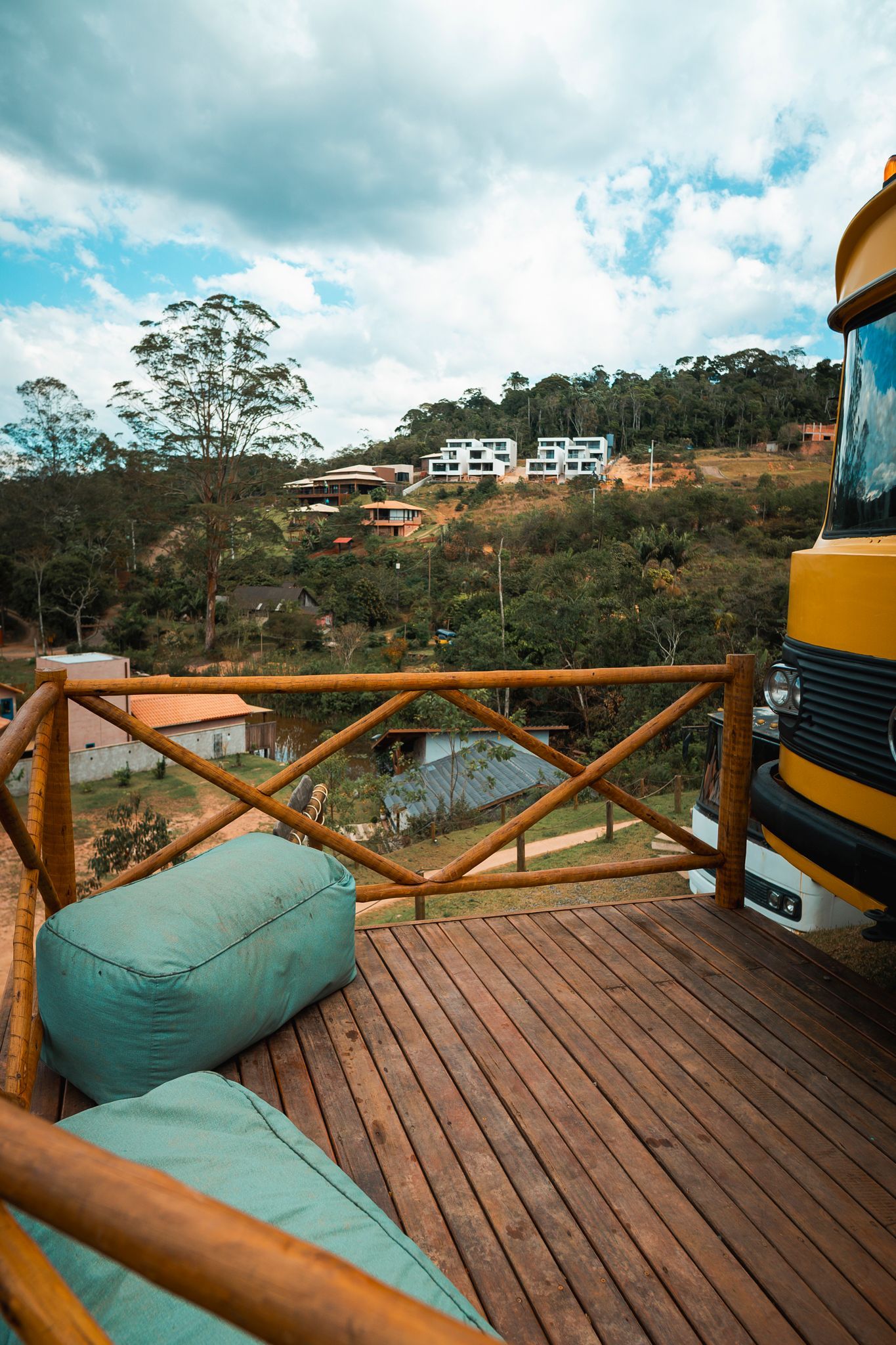 Deck de madeira com pufes e vista parcial de um ônibus amarelo com vista para uma paisagem montanhosa com casas sob um céu nublado. Villa Caravaggio