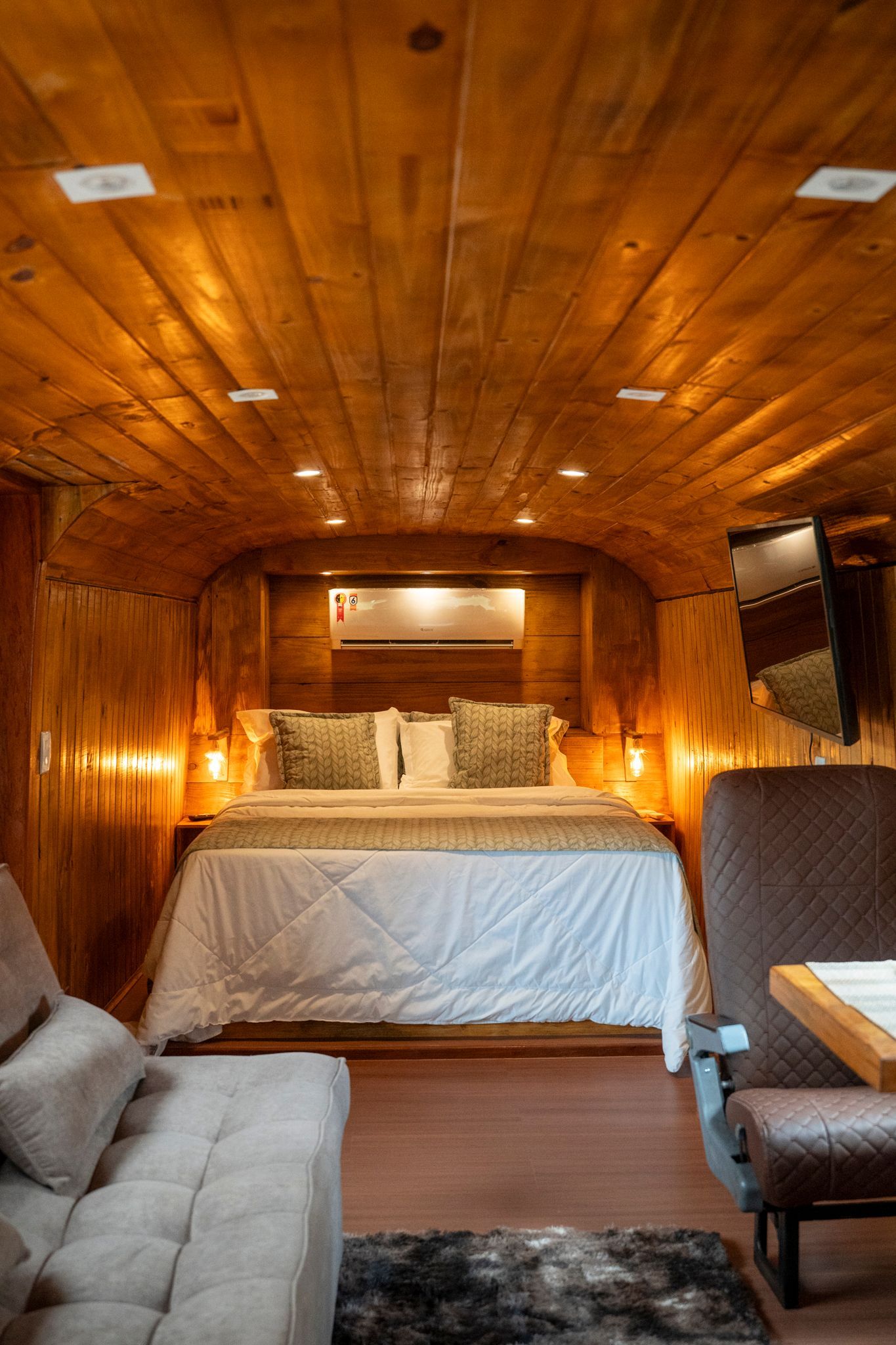 Quarto com paredes de madeira em um ônibus adaptado, com cama, sofá pequeno e TV de parede. Villa Caravaggio