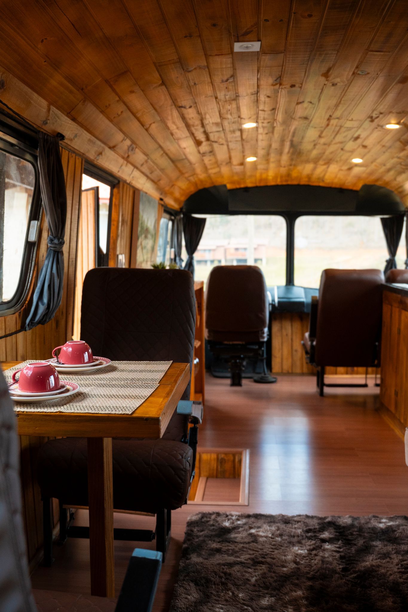 Vista interior de um ônibus convertido em espaço habitável, com detalhes em madeira, uma mesa com xícaras de chá e assentos de motorista. Villa Caravaggio