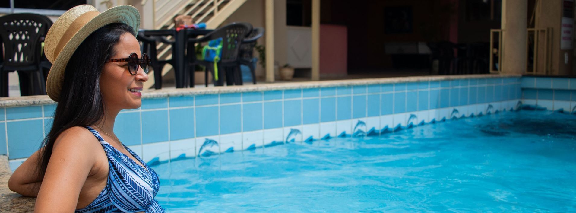 Uma mulher está sentada ao lado de uma piscina usando um chapéu e óculos de sol.