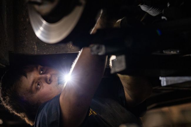 A service mechanic is working under a car with a flashlight.