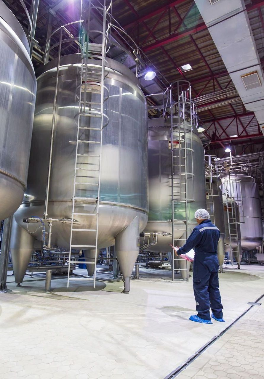 A man is standing in front of a row of stainless steel tanks in a factory.