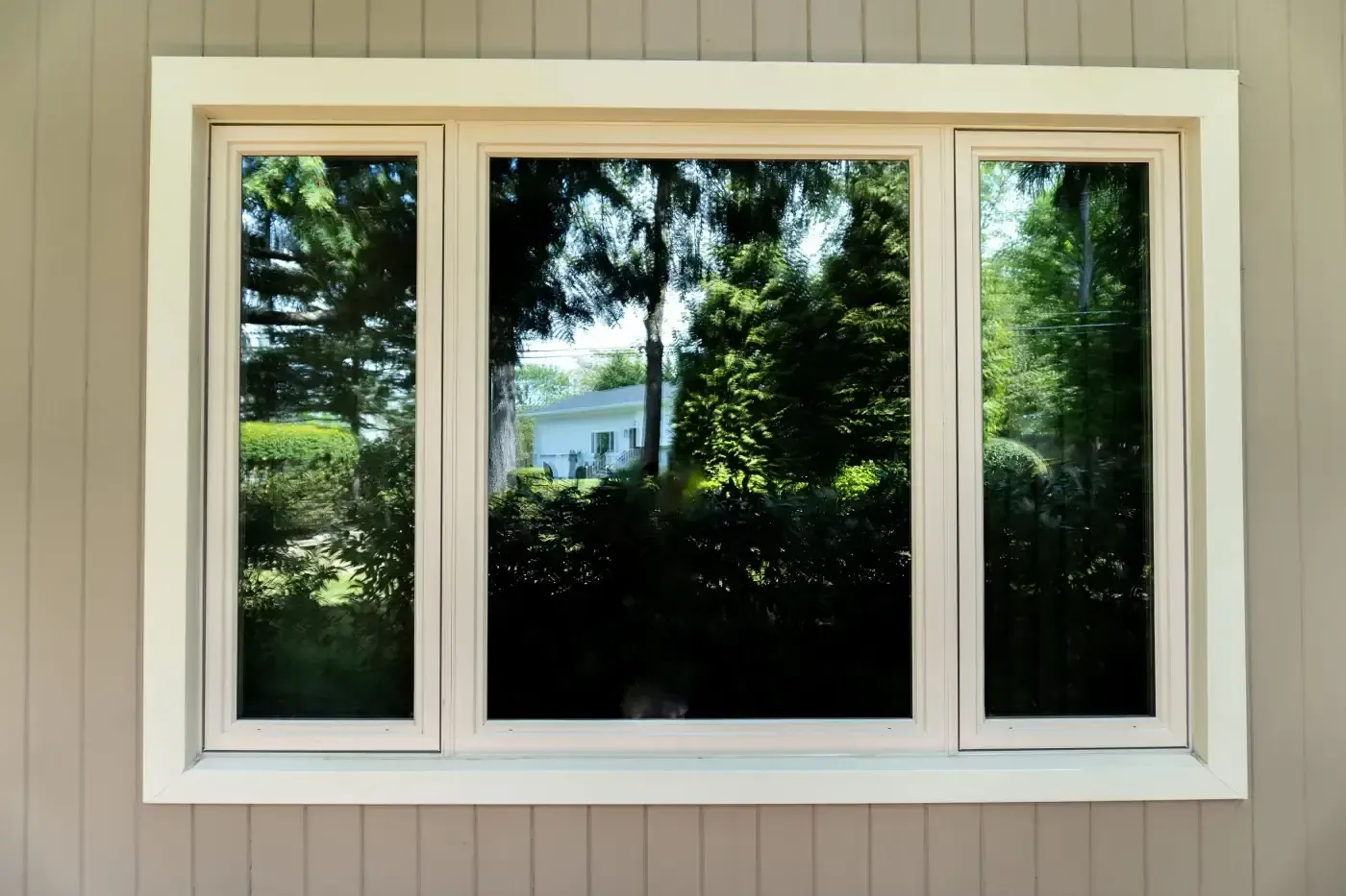White-framed window reflecting green trees and a white house on a light brown siding.