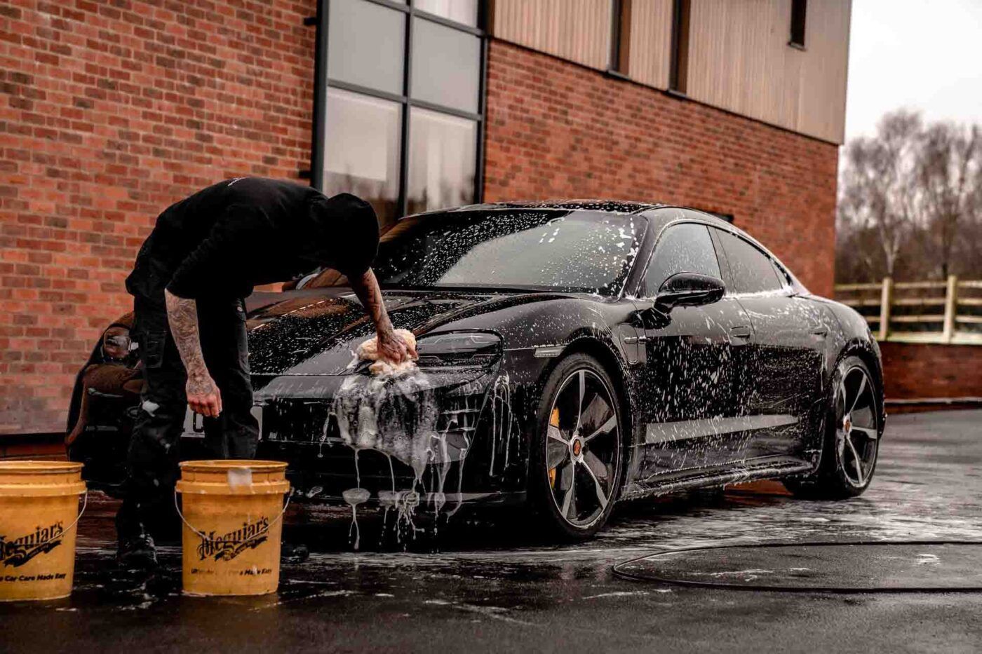 Tattooed male Car detailer washing a Black Porsche Panamera with the two-bucket method with yellow Meguiars 5-gallon buckets
