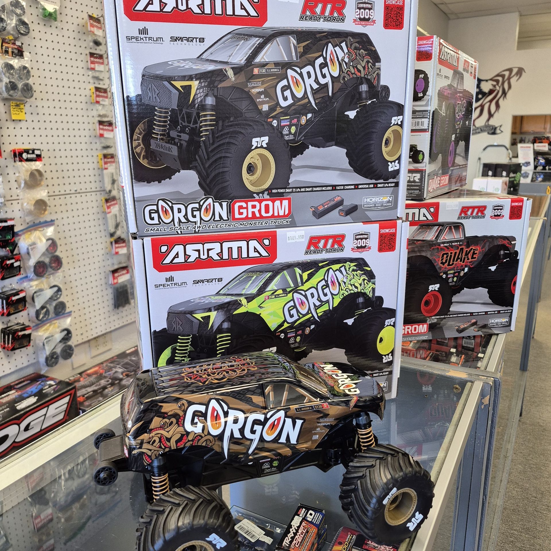 RC monster trucks displayed in a hobby shop. Boxes stacked, brown and green camouflage, black tires.