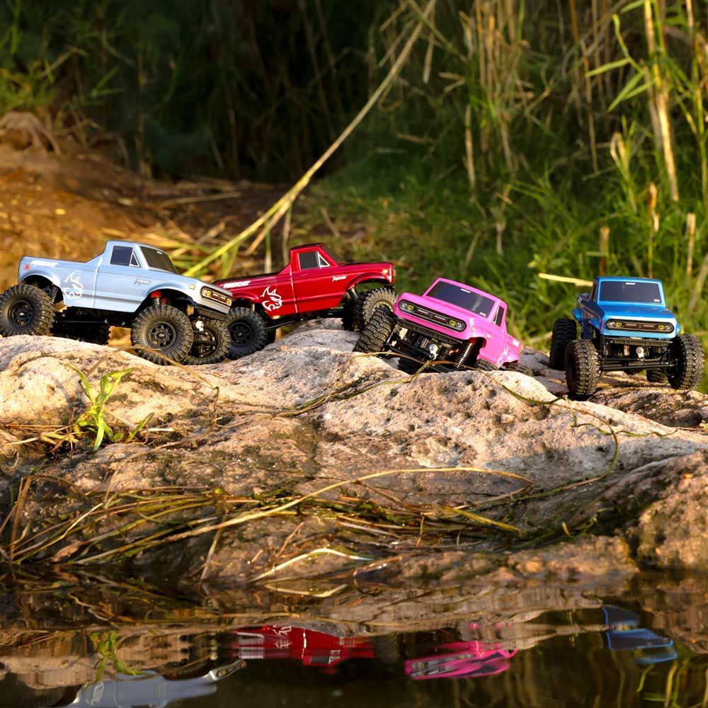 Four colorful miniature off-road trucks on a rocky shore next to a body of water.